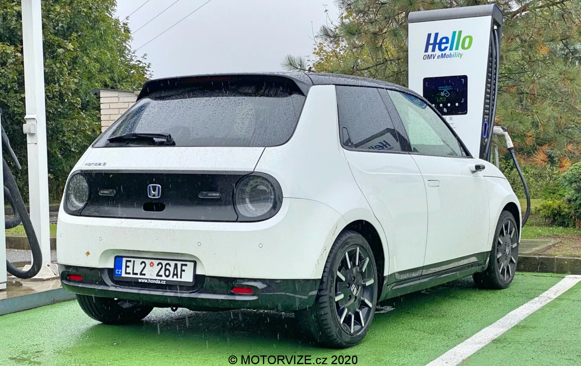 Samochód elektryczny Honda e 2020 sfotografowany z tylnego kąta trzech czwartych, ukazujący prawy tylny ćwiartkowy widok, boczny widok częściowy i tył. Samochód w dwukolorowej kolorystyce, z białą karoserią i czarnym dachem, zaparkowany przy stacji ładowania z podłączonym kablem ładującym z tyłu. Design pojazdu obejmuje okrągłe tylne światła, dużą tylną wycieraczkę, czujniki parkowania, spoiler dachowy i czarne, pięcioramienne felgi aluminiowe. Zdjęcie podkreśla również prostokątną, retro-nowoczesną estetykę samochodu, wykonane w ciągu dnia przy pochmurnej lub deszczowej pogodzie, z samochodem zaparkowanym na specjalnie wyznaczonym miejscu do ładowania samochodów elektrycznych oznaczonym zieloną farbą.