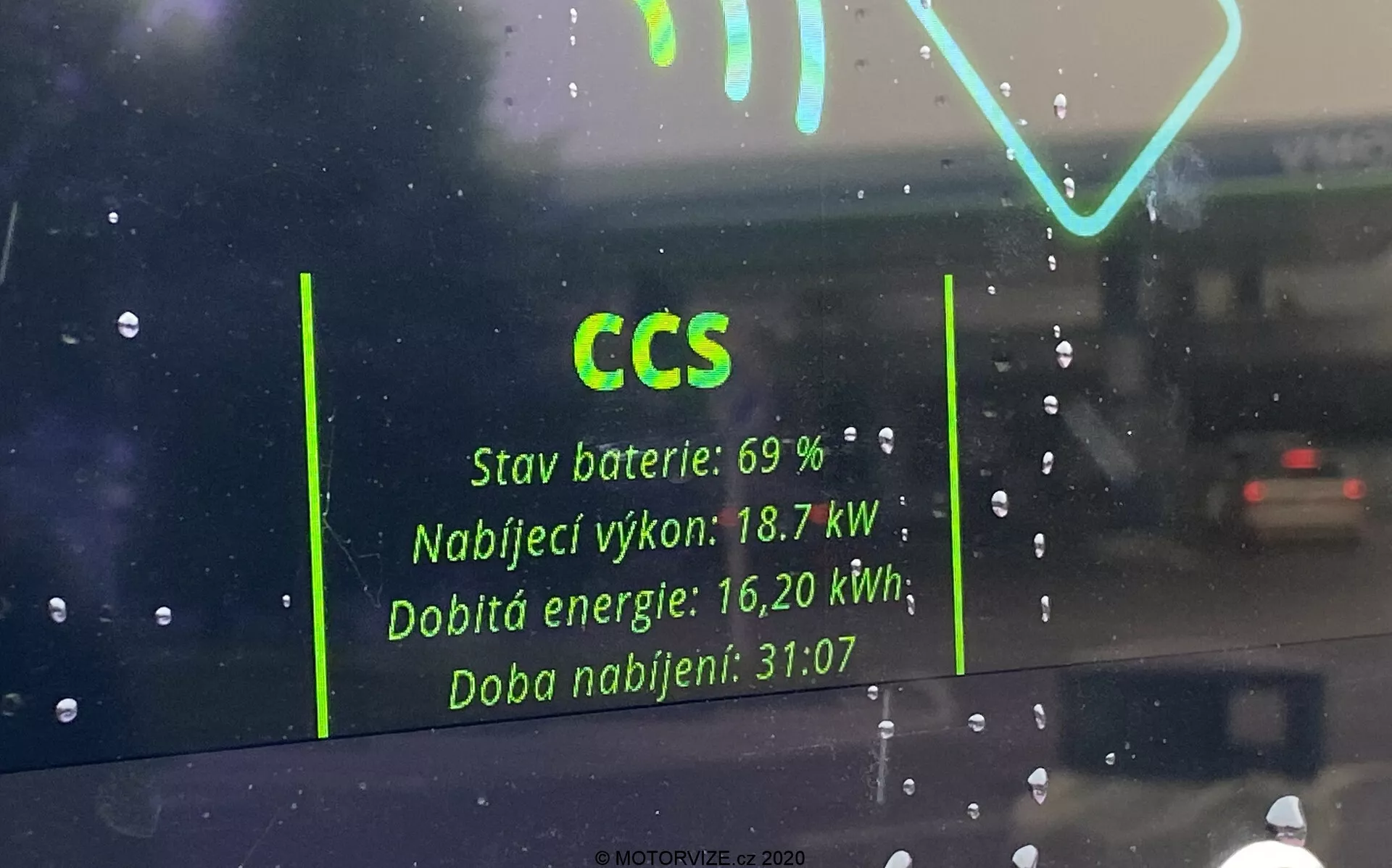 Widok wnętrza przez przednią szybę pojazdu elektrycznego, skupiający się na odbiciu interfejsu stacji ładowania na szkle. Kluczowe szczegóły obejmują 'CCS' dla Combined Charging System, 'Stan baterii: 69 %,' 'Moc ładowania: 18,7 kW,' 'Doładowana energia: 16,20 kWh,' i 'Czas ładowania: 31:07.' Odbicie pojawia się jako zjawa na deszczowym tle z innym samochodem widocznym w oddali.