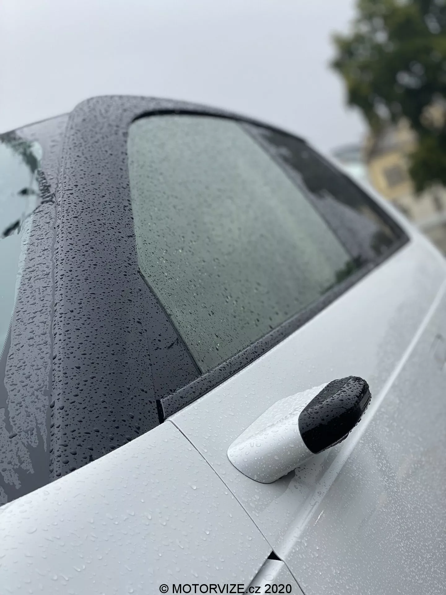 Zbliżenie, kątowy widok tylnej części Hondy e 2020, podkreślający wygląd tylnej bocznej szyby pasażera z kroplami wody oraz jasne tylne drzwi z czarną, wysuwaną klamką, podkreślając zaawansowaną powłokę lakierniczą i system bezkluczykowego dostępu.