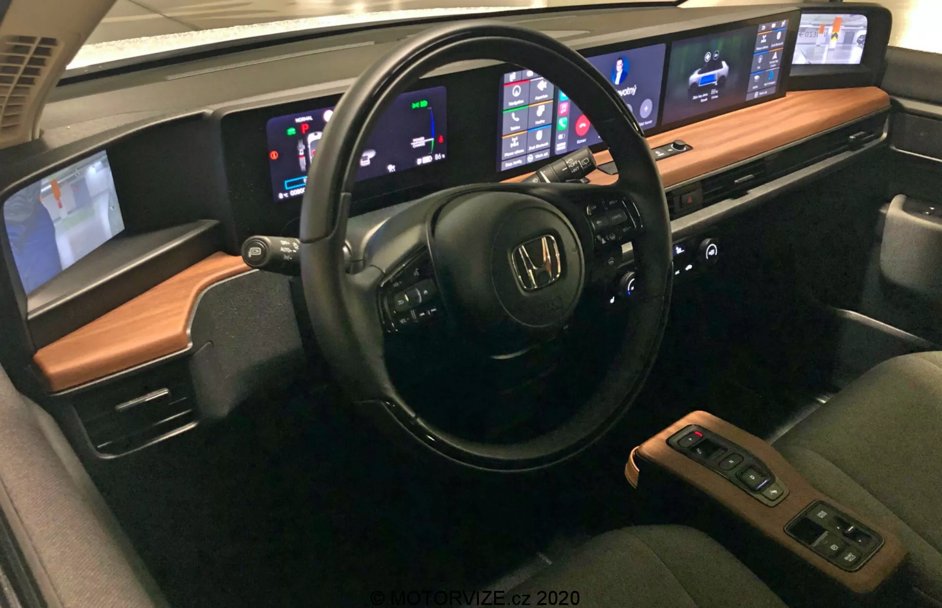 Wnętrze Hondy e 2020 z zaawansowanymi funkcjami, w tym wielofunkcyjną kierownicą z kontrolkami dźwięku i głosu, cyfrowym panelem deski rozdzielczej pokazującym prędkość i poziom paliwa, centralnym systemem infotainment kompatybilnym z Apple CarPlay i Android Auto, automatycznym dwustrefowym systemem klimatyzacji, wygodnymi siedzeniami z materiałem skórzanym i funkcjami grzewczymi, centralną konsolą z dźwignią zmiany biegów i schowkiem, zaawansowanymi systemami wspomagania kierowcy, opcjami łączności jak porty USB i ładowanie bezprzewodowe, wysokiej jakości systemem dźwiękowym oraz konfigurowalnym oświetleniem ambientowym.