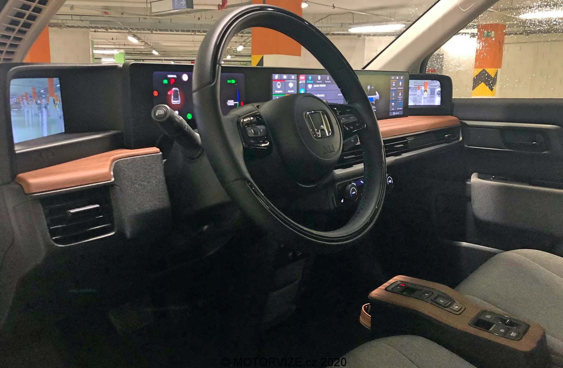 Widok wnętrza Hondy e 2020, skoncentrowany na obszarze kierowcy widzianym z centrum pojazdu. Deska rozdzielcza prezentuje nowoczesny design z dwukolorowym schematem. Kierownica z logo Honda i kilkoma przyciskami sterującymi znajduje się pośrodku, otoczona cyfrowym wyświetlaczem zamiast analogowych wskaźników. Po prawej stronie cyfrowa konsola centralna zawiera panele dotykowe do audio, nawigacji, klimatyzacji i innych ustawień. Design podkreśla integrację technologii, minimalizując przyciski fizyczne na rzecz interfejsów dotykowych. Samochód wydaje się stać na parkingu, z widocznymi przez okna fluorescencyjnymi światłami i betonowymi filarami.