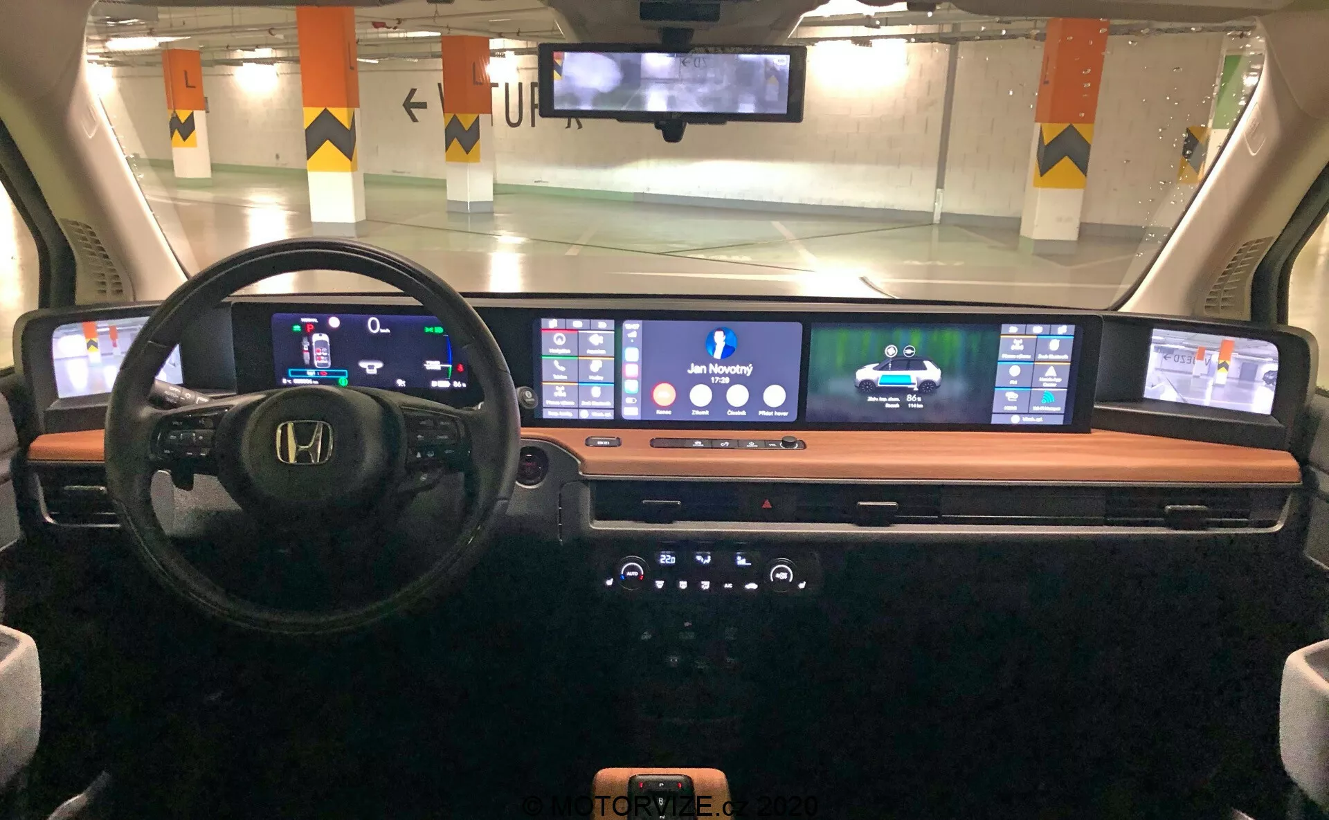 Widok wnętrza deski rozdzielczej Honda e 2020 z perspektywy kierowcy, prezentujący cyfrowy zestaw wskaźników, wielofunkcyjną kierownicę, rozległy ciągły ekran cyfrowy do nawigacji i mediów, elegancką konsolę centralną z przyciskami i pokrętłami oraz bezprzewodową ładowarkę do telefonu, podkreślając nowoczesną technologię motoryzacyjną i design.