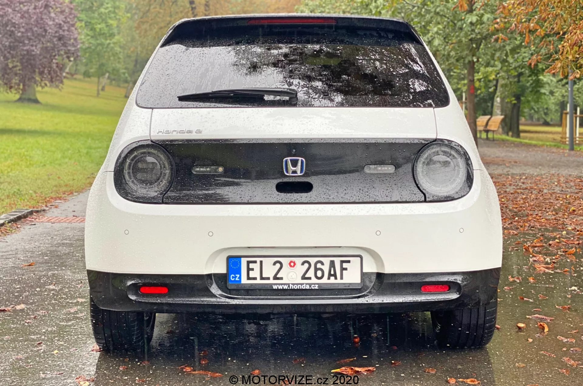 Widok tylnej części białego, mokrego samochodu Honda e 2020, ukazujący okrągłe tylne światła LED, trzecie światło hamowania, emblemat Honda, tylna klapa z wycieraczką, europejska tablica rejestracyjna, czujniki parkowania, tylne światła przeciwmgielne, tylne spojler oraz elementy odblaskowe na zderzaku. Zdjęcie uwiecznia detale takie jak krople wody na pojeździe, z rozmytym, parkowym tłem z drzewami i trawą.