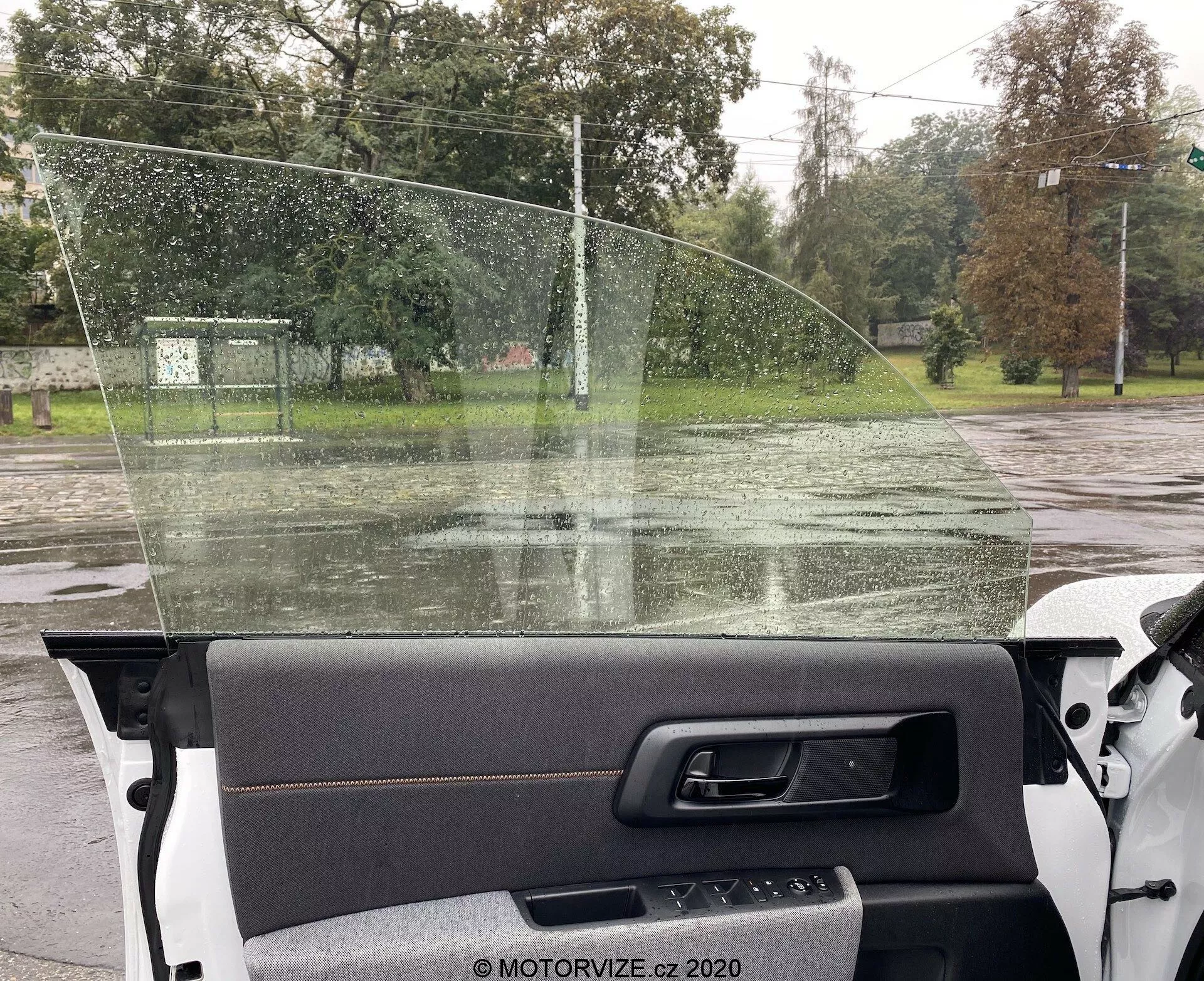 Widok wnętrza z perspektywy pasażera w Honda e 2020, ukazujący prawy panel drzwiowy z wstawką z tkaniny i metaliczną klamką, przednią szybę z kroplami deszczu oraz widok na mokrą drogę z drzewami i ogrodzeniem za nią.