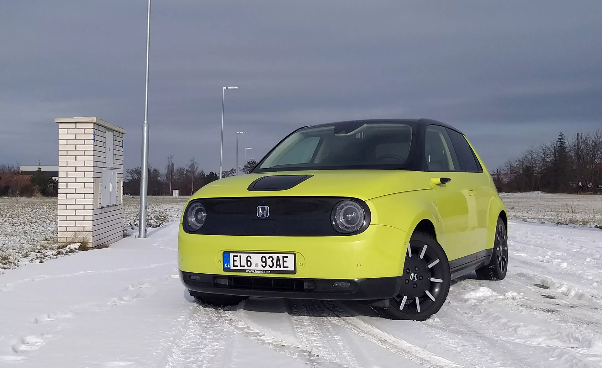 Honda e 2020 w jasnożółtym kolorze na powierzchni pokrytej śniegiem z pochmurnym niebem, z reflektorami LED, technologią pojazdu elektrycznego z zaawansowanymi systemami wspomagania kierowcy, czarnymi alufelgami i oponami zimowymi.