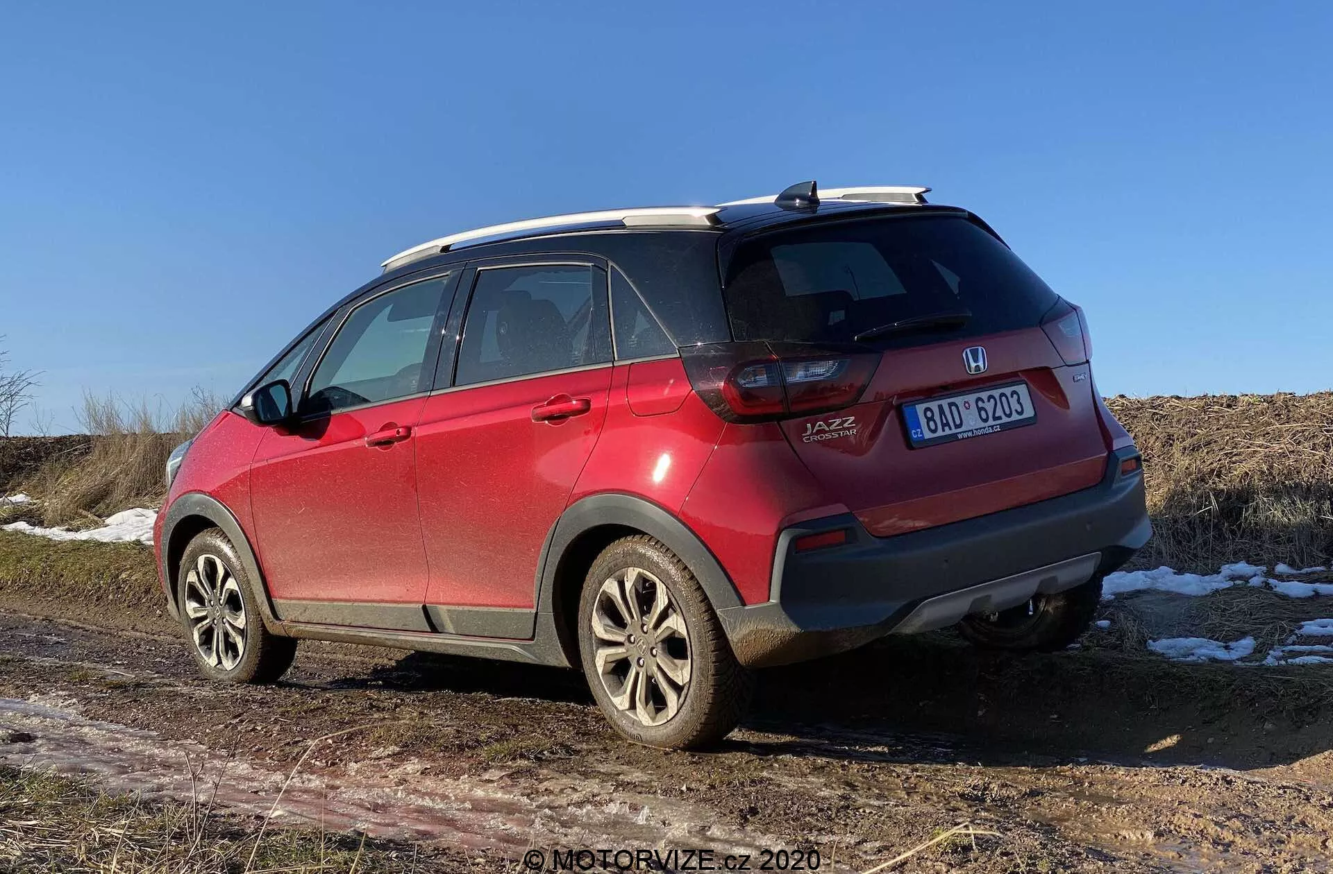 Czerwony SUV Honda Jazz Crosstar 2020 z widokiem z trzech czwartych tyłu, ukazujący szczegóły takie jak emblematy marki i modelu, tylne światła LED, tylny zderzak z rysami i czujnikami, aluminiowe felgi, opony całoroczne, relingi dachowe, antena typu płetwa rekina, przyciemniane szyby z liniami odmrażającymi i centralną tablicę rejestracyjną. Otwieranie bagażnika typu hatchback i plamy śniegu na ziemi z suchym polem w tle sugerują późną jesień, zimę lub wczesną wiosnę przy czystym niebie w południe lub wczesnym popołudniem.