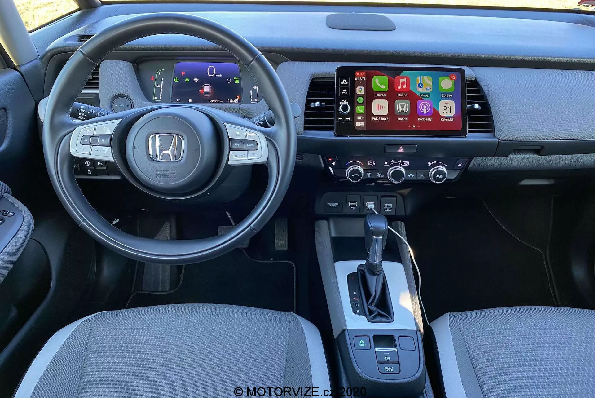 Widok wnętrza Honda Jazz Crosstar 2020 z perspektywy kierowcy. Zmodernizowana, wielofunkcyjna kierownica z widocznym logo Honda, posiadająca przyciski do obsługi audio, tempomatu i komend głosowych. Za kierownicą znajduje się cyfrowy panel instrumentów wyświetlający kluczowe informacje. Konsola centralna zawiera system informacyjno-rozrywkowy wspierający prawdopodobnie Apple CarPlay lub Android Auto, a poniżej przyciski kontrolne klimatyzacji. Prosta dźwignia zmiany biegów oraz dodatkowe przyciski do obsługi hamulca postojowego i wyboru trybu jazdy są również widoczne. Oświetlenie i kąt ujęcia podkreślają wygodę i ergonomiczne rozmieszczenie elementów.