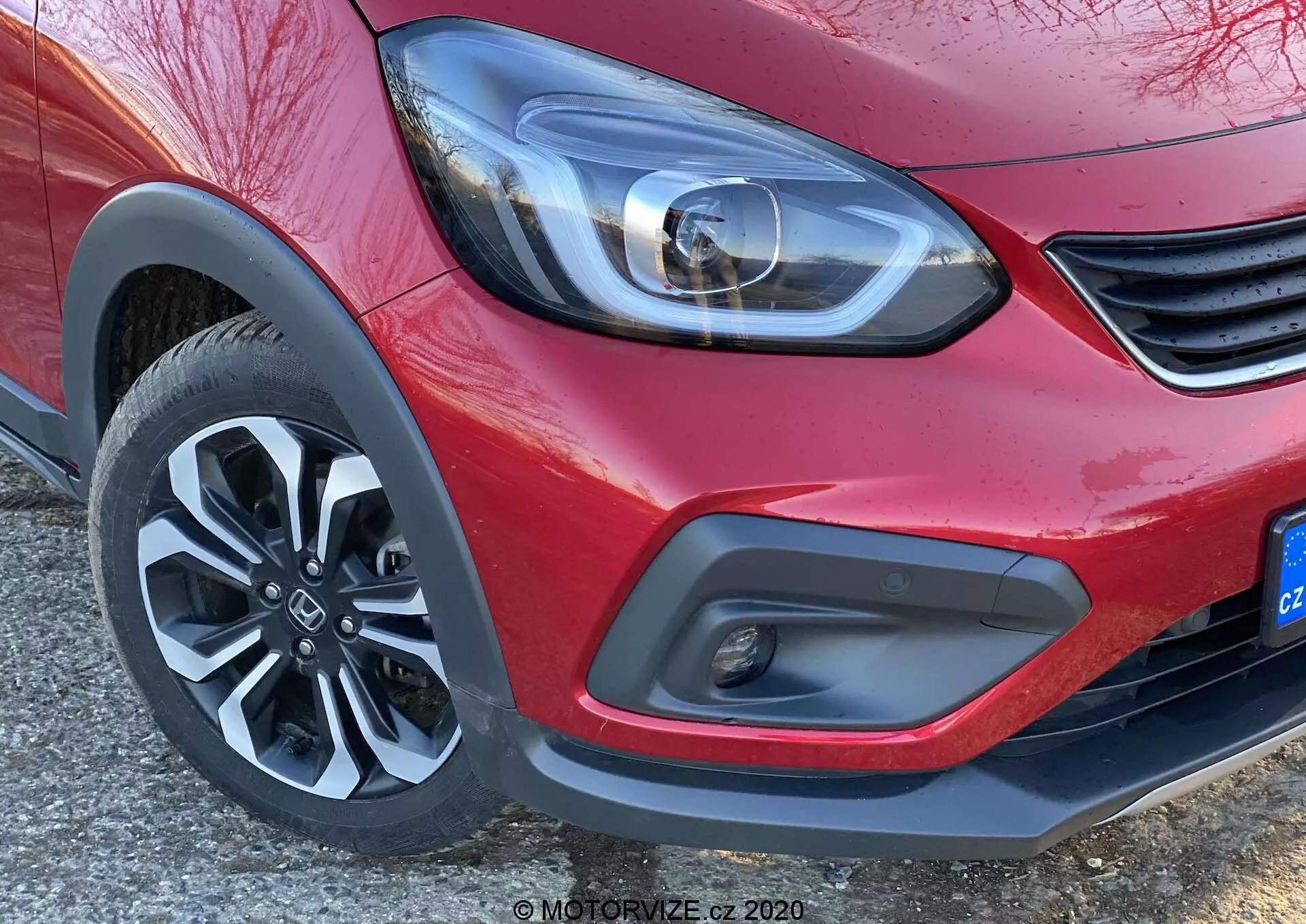Zbliżenie na przedni narożnik czerwonej Hondy Jazz 2020 (Jazz Crosstar), skupiające się na nowoczesnym reflektorze LED, minimalistycznym detalu grilla, dwukolorowym przednim zderzaku z reflektorem przeciwmgielnym i dynamicznym pięcioramiennym kole aluminiowym.