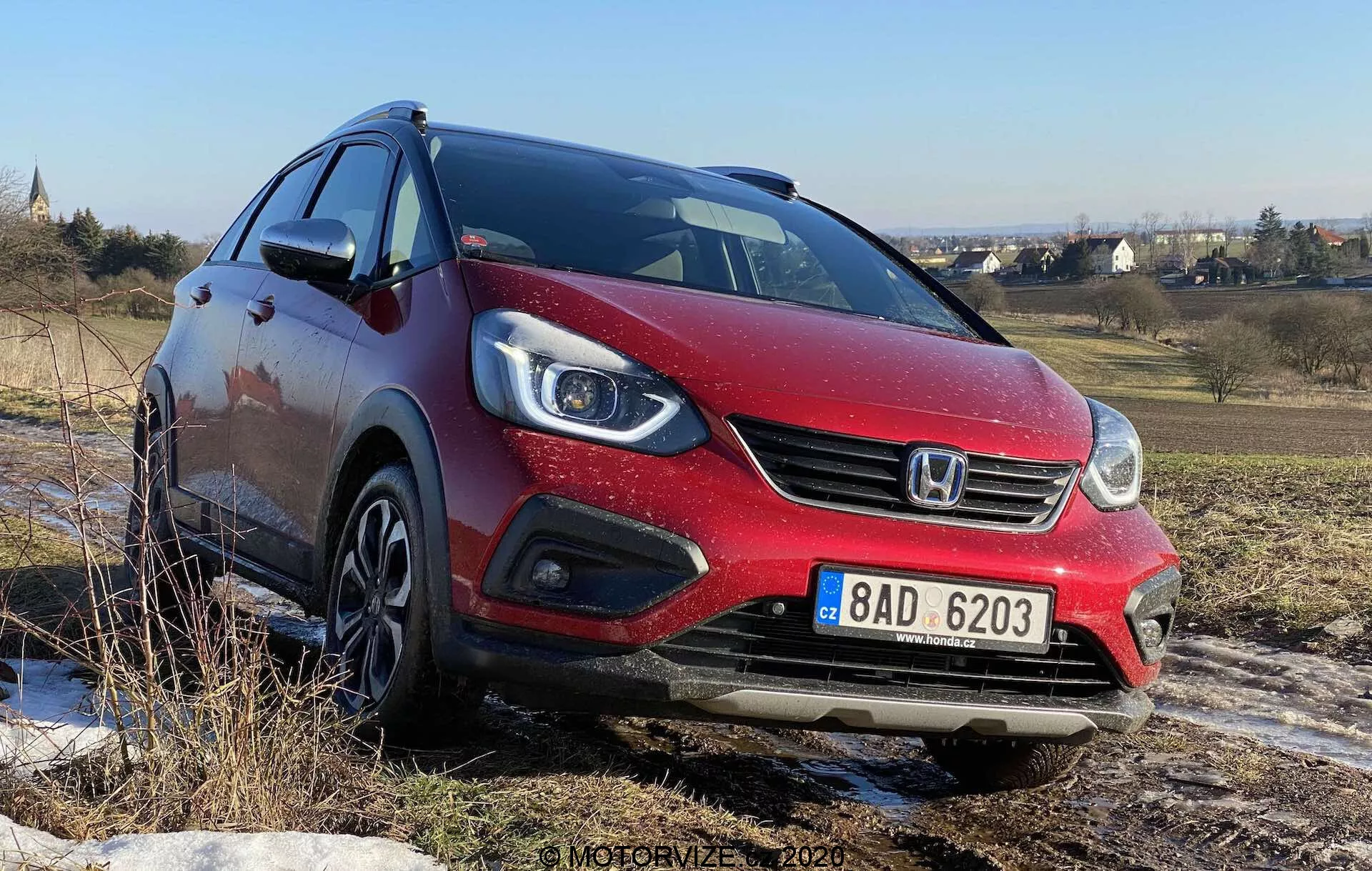 Trzy-czwarta widok przedni czerwonego hatchbacka Honda Jazz Crosstar 2020, uchwycony z niskiego kąta, ukazujący jego przednią fasadę, halogenowe reflektory projektorowe z zintegrowanymi światłami do jazdy dziennej LED, przedni grill z logo marki oraz felgi aluminiowe. Samochód znajduje się na nierównym terenie pod czystym niebieskim niebem, sugerując jego zdolność do poruszania się po nieutwardzonych nawierzchniach.