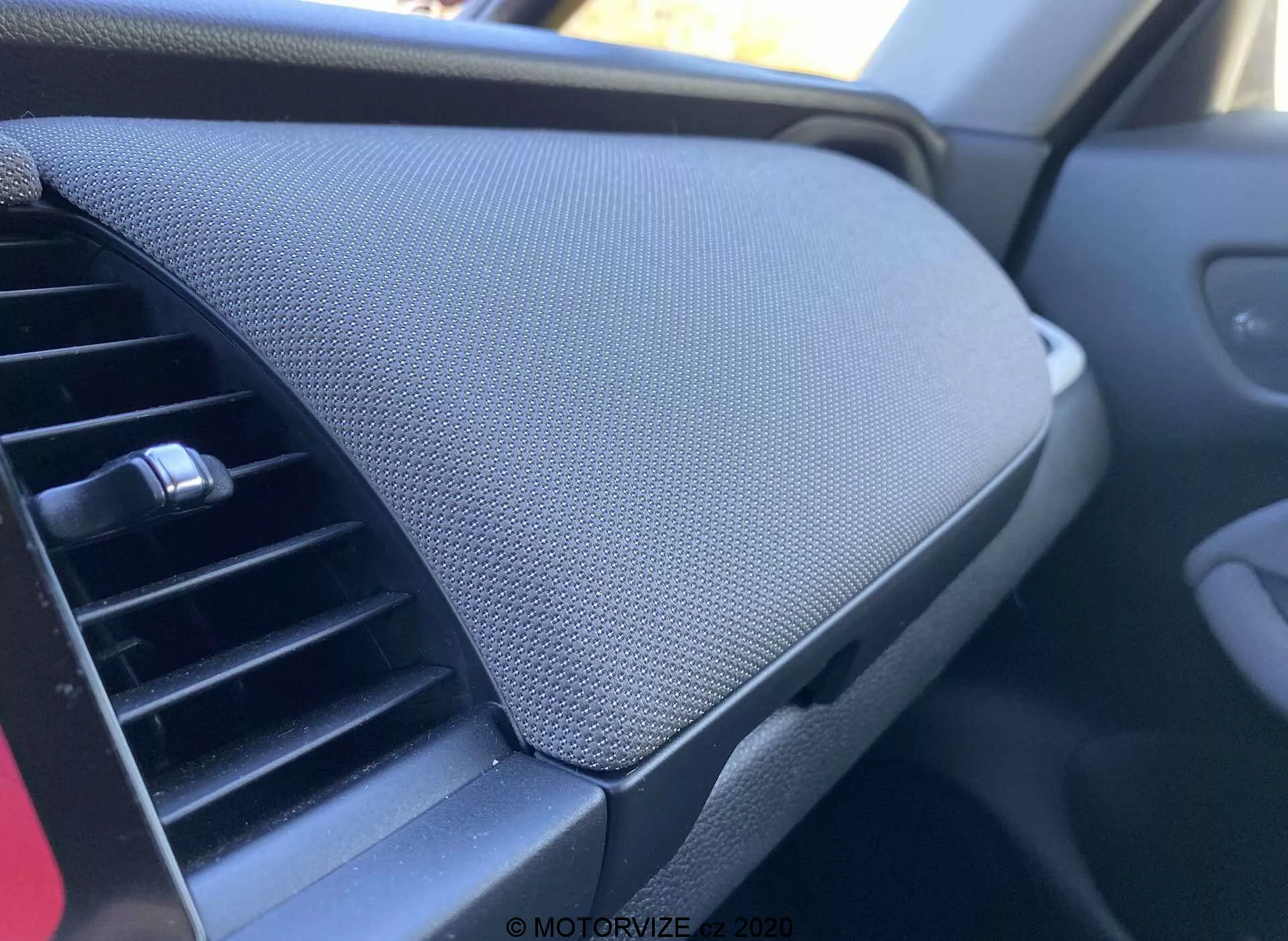 Zbliżony widok wnętrza samochodu Honda Jazz 2020 (Jazz Crosstar), koncentrujący się na desce rozdzielczej. Zdjęcie zrobione z perspektywy pasażera pokazuje wentylację po lewej stronie, z regulowanym poziomo wzorem. Po prawej stronie zdjęcia dominuje schowek na desce rozdzielczej z zamkniętą pokrywą, charakteryzowany fakturowaną powierzchnią, prawdopodobnie z siatki lub tkaniny. Schowek jest płynnie zintegrowany bez widocznej rączki, co sugeruje mechanizm otwierania na przycisk. Ogólny schemat kolorów jest ciemny, co sugeruje neutralną estetykę ukierunkowaną na zmniejszenie rozproszenia uwagi.