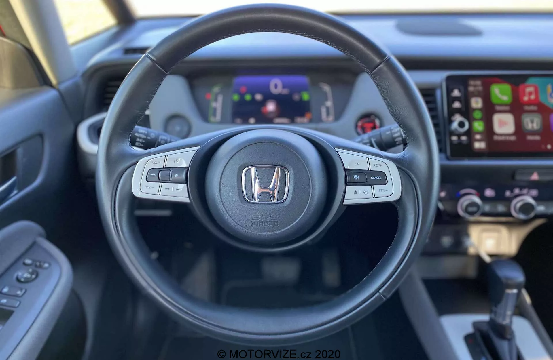 Wnętrze Honda Jazz Crosstar 2020 widziane z fotela kierowcy, ukazujące kierownicę z zamontowanymi przyciskami do sterowania audio, głosowego i tempomatu. Emblemat Honda jest wyeksponowany w centrum. Za kierownicą znajduje się cyfrowy zestaw wskaźników pokazujący różne mierniki i informacje. Po prawej widoczny jest ekran dotykowy systemu infotainment z funkcją Apple CarPlay, a także lekko rozmyte kontrolki klimatyzacji. Zdjęcie wykonane z perspektywy pierwszej osoby podkreśla ergonomiczny design i dostępność technologii pojazdu.
