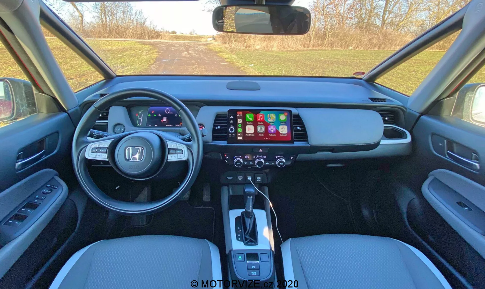 Widok wnętrza nowoczesnego samochodu Honda Jazz 2020 (Jazz Crosstar) z perspektywy tylnych siedzeń, przedstawiający przednią deskę rozdzielczą z cyfrowym panelem instrumentów, dużym dotykowym ekranem systemu informacyjno-rozrywkowego, panelem sterowania klimatyzacją, automatyczną dźwignią zmiany biegów, skórzanymi siedzeniami i przyciskami sterowania na drzwiach.