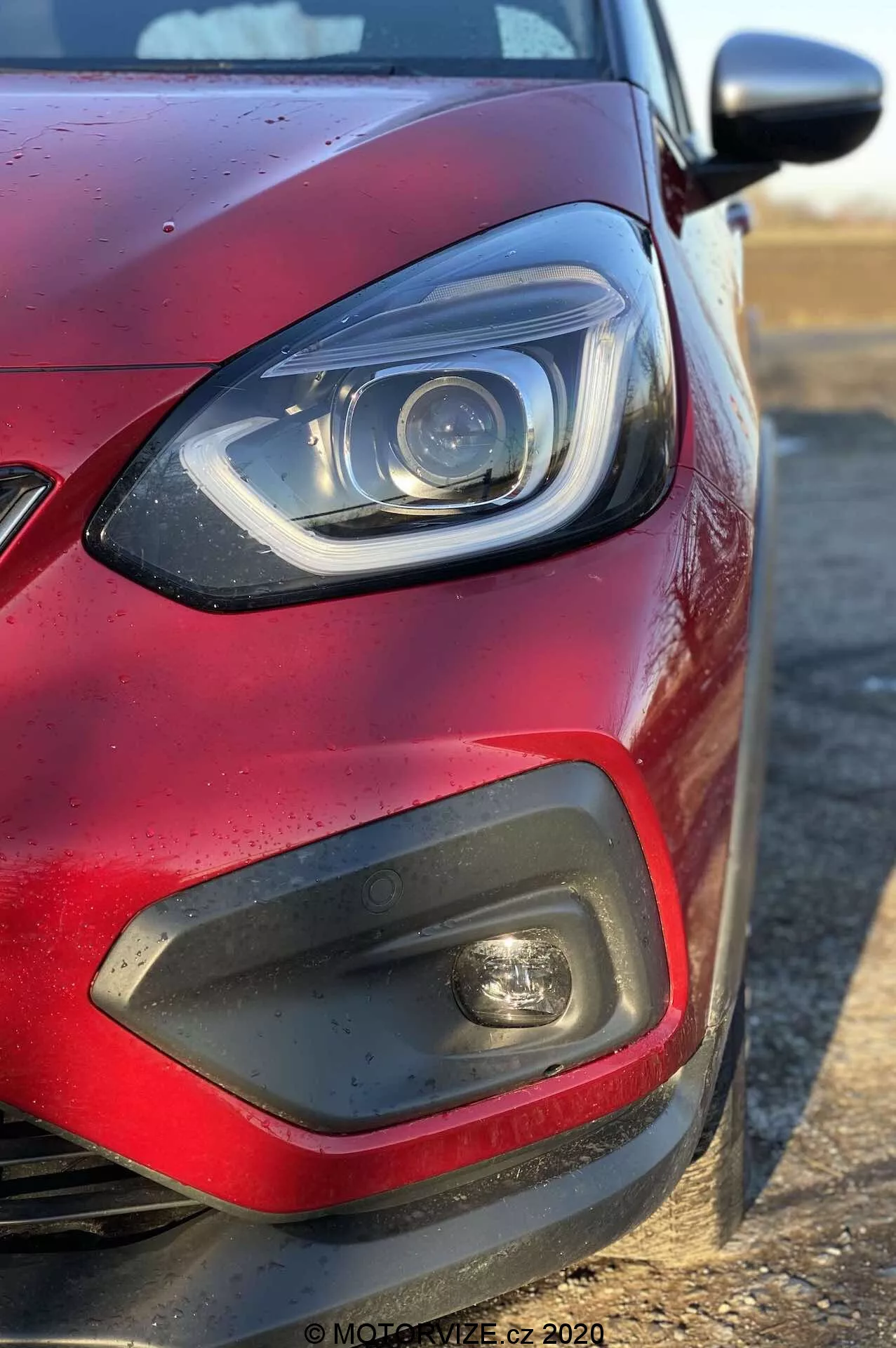 Zbliżenie przedniego lewej kwadrantu czerwonego Honda Jazz 2020 (Jazz-Crosstar) z bocznego kąta, skupiające się na reflektorze z elementami LED lub ksenonowymi i zakrzywioną listwą DRL, czarnym plastikowym wstawce z światłem przeciwmgielnym, części grilla, metalicznym czerwonym lakierze z kroplami wody i lusterku bocznym w kolorze nadwozia wysuwającym się z prawej strony.