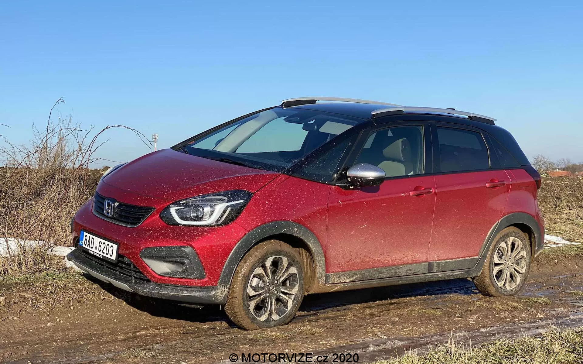 Widok trzy czwarte przód czerwonego SUV-a crossover Honda Jazz Crosstar 2020, uchwyconego w świetle dziennym od strony pasażera, pokazujący przednią kratkę, reflektory, felgi ze stopów pokryte błotem i profil boczny do tylnych drzwi pasażera. Samochód wydaje się lekko zabrudzony, co wskazuje na niedawne użycie na nieutwardzonych lub mokrych drogach. Widoczne cechy to wyraźna kratka z logo marki, nowoczesne reflektory LED, światła przeciwmgielne, felgi ze stopów, lusterka boczne z zintegrowanymi kierunkowskazami, relingi dachowe i podwyższony prześwit, sugerujące wszechstronność i zdolność pojazdu do lekkiej jazdy terenowej. SUV jest zaparkowany w wiejskiej scenerii z suchą trawą i czystym niebem, podkreślając jego przydatność do wiejskich dróg i lekkiej jazdy terenowej.