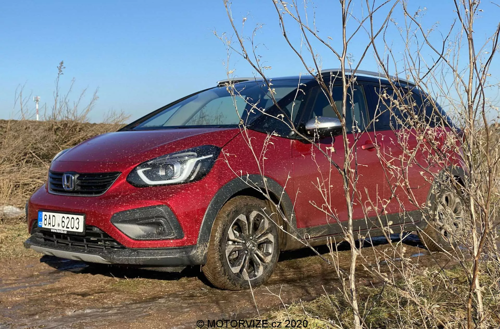 Dzienna fotografia zewnętrzna Hondy Jazz 2020 (Jazz Crosstar) zaparkowanej na trawiastej powierzchni. Obraz pokazuje czerwony kolor subkompaktowego SUV-a, wyraźny grill oraz nowoczesne reflektory z oświetleniem LED. Samochód widziany jest pod kątem trzech czwartych przodu, ukazując zwiększoną wysokość pojazdu, aluminiowe felgi oraz czarne plastikowe osłony wokół łuków kół i dolnych paneli karoserii. Jasne niebo, naturalne oświetlenie i otaczające gałęzie częściowo zasłaniają dolną przednią część. Widoczne są lusterka boczne z zintegrowanymi kierunkowskazami oraz widok wnętrza przez przezroczystą szybę po stronie kierowcy.