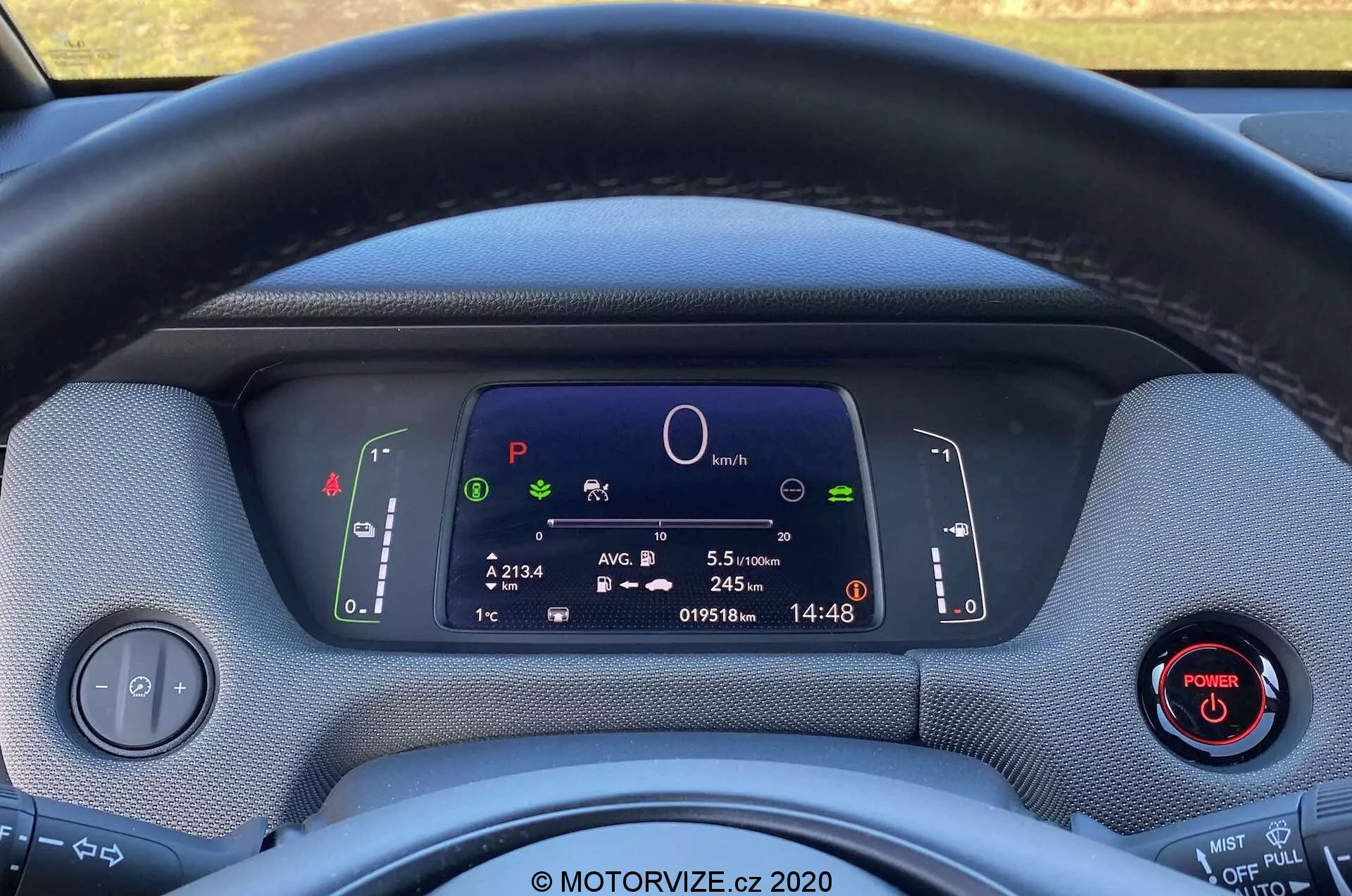 Zbliżenie wnętrza Honda Jazz Crosstar 2020 z perspektywy kierowcy, ukazujące kierownicę i w pełni cyfrowy zestaw wskaźników. Kierownica wykonana z tkaniny częściowo zasłania widok na cyfrowy ekran, na którym widać symbole systemów auta, centralny okrągły wykres, informacje o podróży, licznik przebiegu i zegar. Dwa dźwignie za kierownicą kontrolują kierunkowskazy, światła i wycieraczki. Panel sterowania światłami i przycisk z czerwonym pierścieniem są widoczne odpowiednio po lewej i prawej stronie. Materiały deski rozdzielczej maja teksturowany wygląd, co podkreśla design ekologiczny. Zdjęcie podkreśla nowoczesną technologię, cyfrową instrumentację i możliwe cechy hybrydowe lub elektryczne pojazdu.