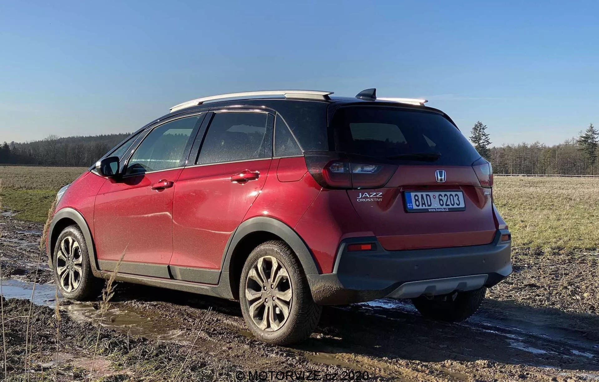 Widok tylnego narożnika czerwonego Honda Jazz Crosstar 2020, ukazujący jego cechy crossover SUV. Zdjęcie wykonane pod niskim kątem podkreśla wysokość pojazdu i prześwit, z widocznymi zespołami świateł tylnych, oznaczeniem 'JAZZ', felgami aluminiowymi, systemem relingów dachowych, przyciemnianymi tylnymi szybami, odpryskami błota i widoczną tablicą rejestracyjną, zaparkowany na błotnistej polanie.