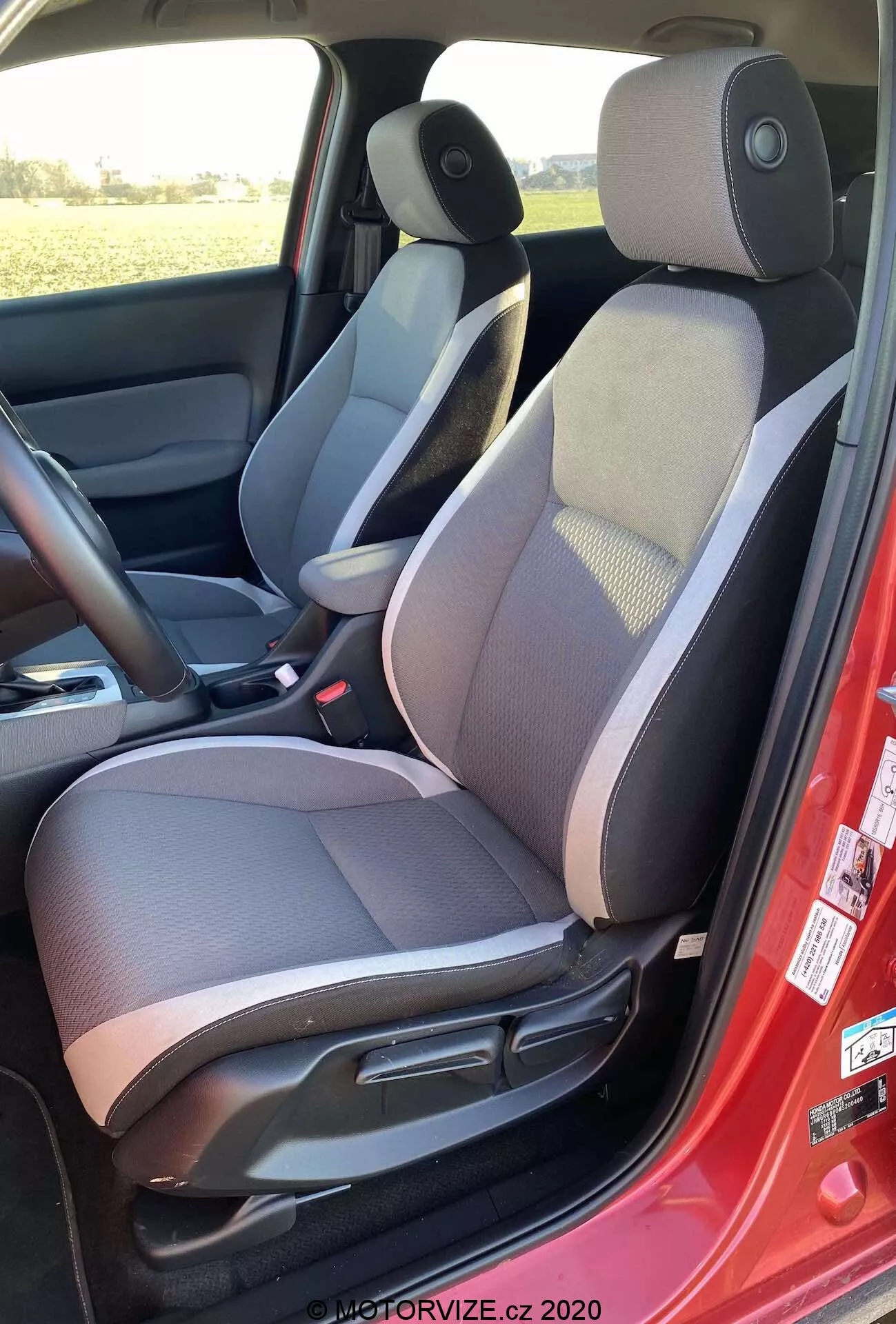 Widok wnętrza Honda Jazz Crosstar 2020 od lewej strony, podkreślający dwukolorową tapicerkę fotela kierowcy z wzorzystą środkową częścią, regulowanym zagłówkiem, manualnymi kontrolkami regulacji fotela, czerwoną pasem bezpieczeństwa, głośnikiem w panelu drzwi, ciemną matą podłogową, centralną konsolą z podłokietnikiem, kontrolką regulacji bocznego lusterka oraz ciemnym i jasnym schematem kolorów.