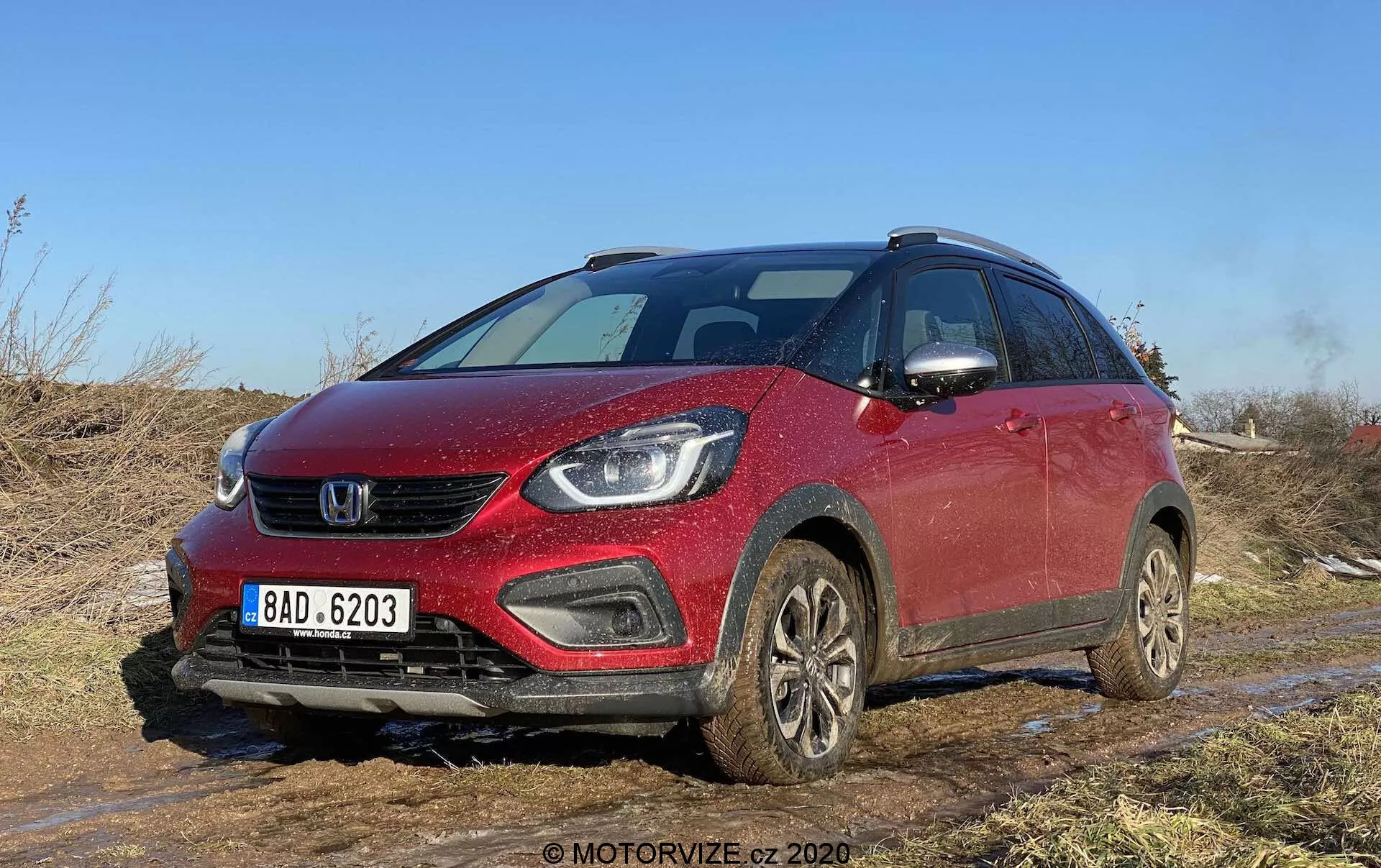 Widok z przodu z kąta na czerwony Honda Jazz Crosstar 2020, kompaktowy crossover SUV, uchwycony na terenie off-road z widocznym błotem i ziemią. Obraz podkreśla przednią kratkę, smukłe reflektory mogące zawierać lampy LED lub projektorowe, zintegrowane w zderzaku światła przeciwmgielne oraz delikatnie opadającą maskę. Pojazd, wyposażony w relingi dachowe, koła pokryte błotem i lusterka boczne z wbudowanymi kierunkowskazami, sugeruje nowoczesne funkcje bezpieczeństwa i zaawansowane wyposażenie technologiczne. Idealny dla kategorii dotyczących pojazdów, recenzji samochodów, zdolności terenowych, technologii motoryzacyjnych i wzornictwa.