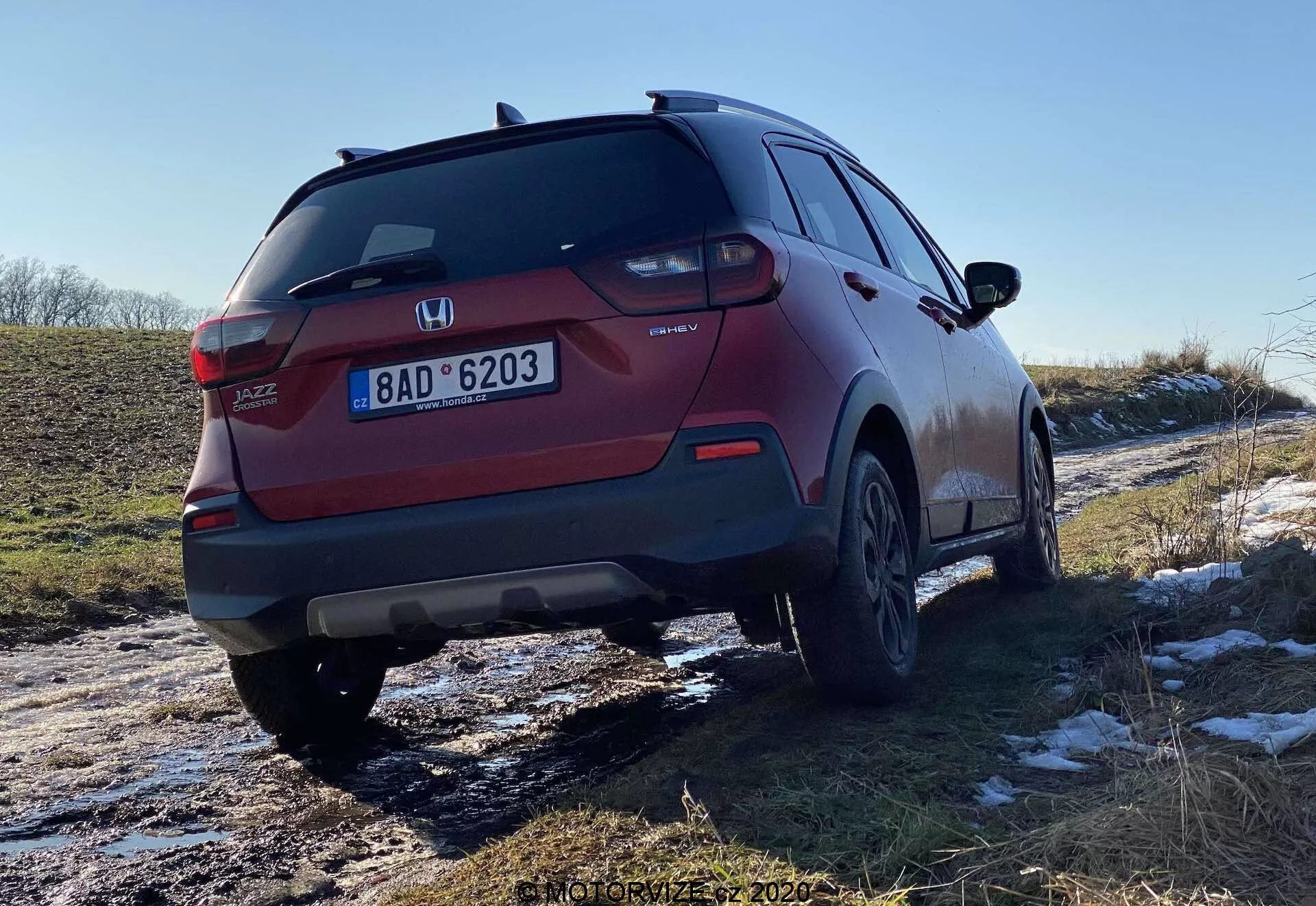 Widok tylno-trzyczwarty czerwonego SUV-a Honda Jazz Crosstar, ukazujący design świateł tylnych, tylnego zderzaka, układu wydechowego i felg aluminiowych. Kąt podkreśla prześwit pojazdu, a ślady śniegu i błota świadczą o jego wszechstronności w różnych warunkach drogowych.