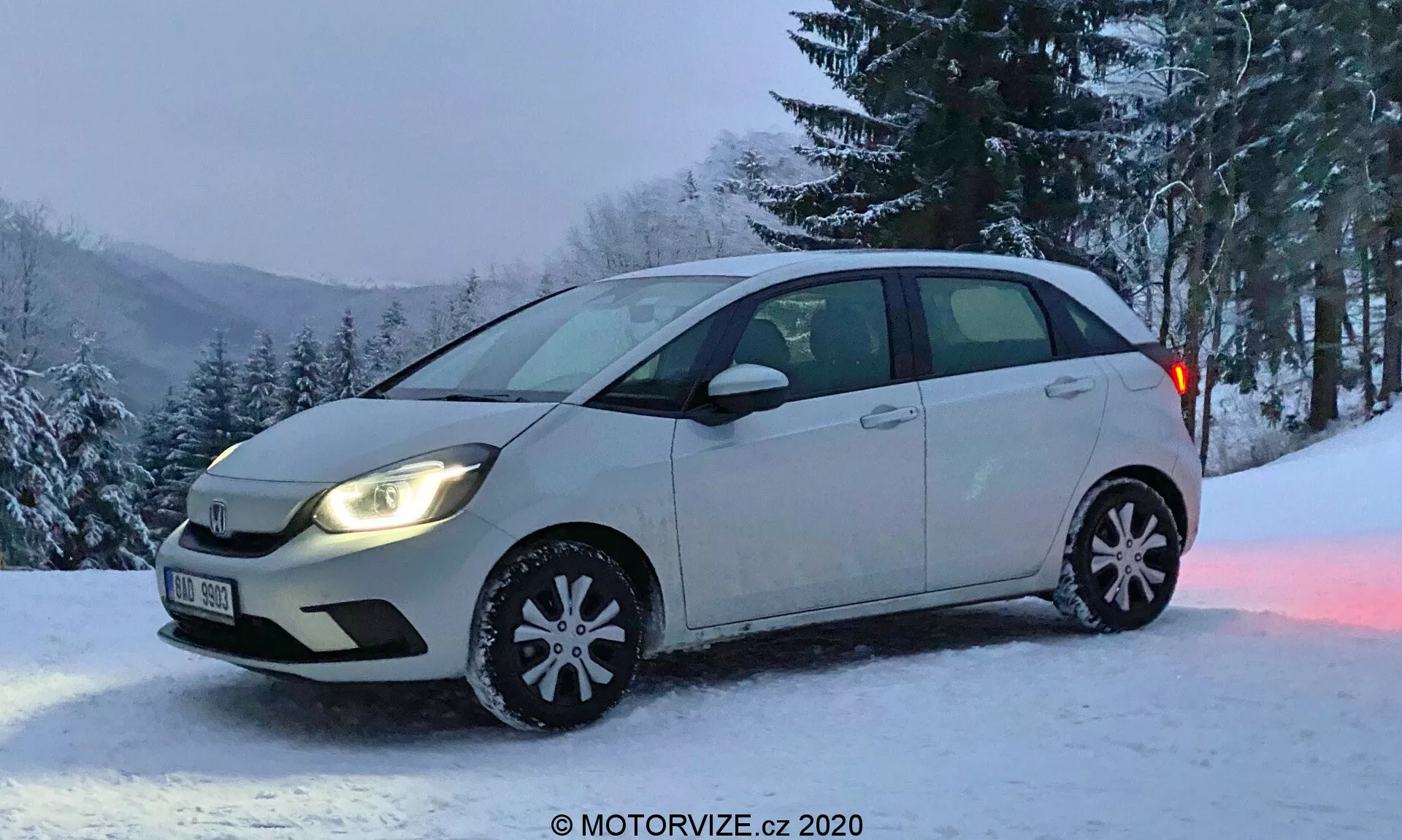 Honda Jazz 2020 hatchback kompaktowy sfotografowany pod ukośnym kątem przednim, ukazujący pełny widok na stronę kierowcy oraz częściowy widok na przedni zderzak i kratkę. Pojazd ma dwukolorowy schemat kolorów nadwozia z większością w jasnym kolorze i dolną częścią drzwi bocznych w ciemniejszym odcieniu. Światła są włączone, z typowymi lampami halogenowymi, bez świateł przeciwmgielnych. Widoczne są dekoracyjne zimowe osłony kół z odblaskowymi płatkami śniegu, wskazując na sezon zimowy. Samochód zaparkowany jest na zaśnieżonym terenie z tłem drzew i zaśnieżonego wzgórza, co sugeruje wiejską lub górzystą lokalizację. Zdjęcie dzięki uprzejmości MOTORVIZE.cz.