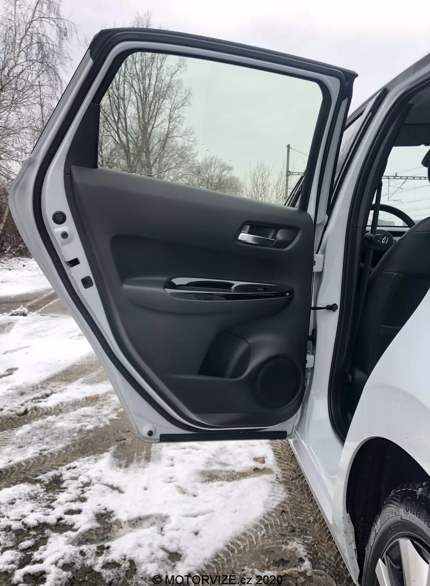 Zbliżenie wnętrza przednich drzwi kierowcy Honda Jazz 2020, ukazujące szczegółowy widok teksturowanego wykończenia z tworzywa sztucznego, ergonomicznej satynowej klamki, przycisków do sterowania szybami i lusterkami elektrycznymi, formowaną kieszeń na przechowywanie i grill głośnika, wszystko to w zaśnieżonym zimowym otoczeniu.