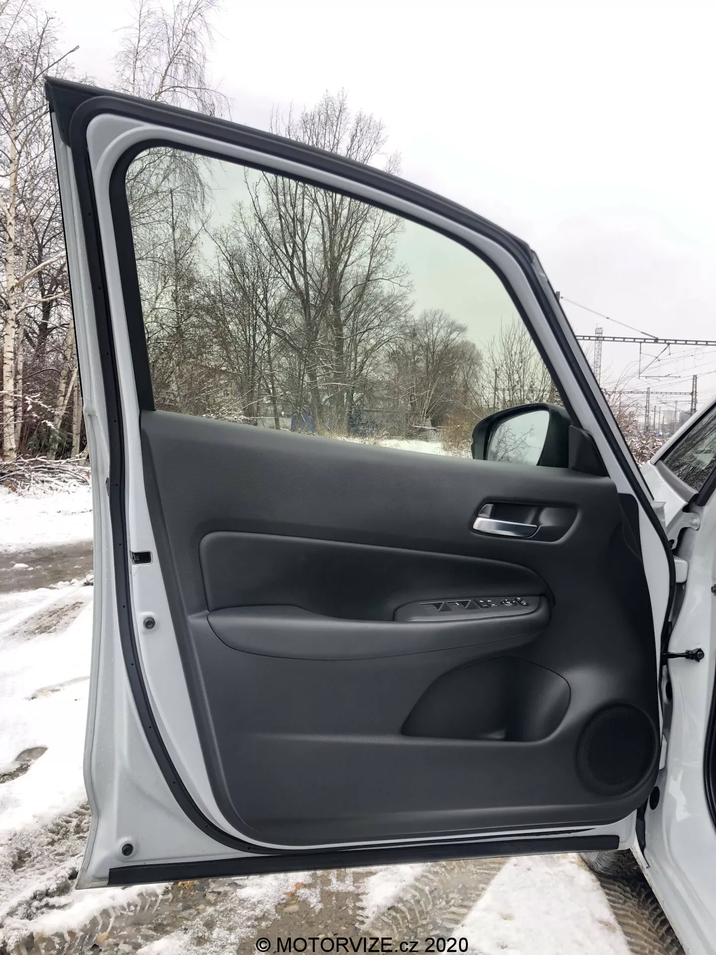 Widok wnętrza przednich drzwi pasażera Honda Jazz 2020, w pełni otwartych, prezentujący teksturowane i gładkie czarne materiały plastikowe. Panel drzwi zawiera ergonomiczny podłokietnik, miękką klamkę i panel kontrolny do elektrycznych mechanizmów okiennych i zamykających. Widoczny jest również srebrny akcent i dolna kieszeń na przechowywanie. Przez okno widać zaśnieżony krajobraz, co sugeruje zimowe otoczenie.
