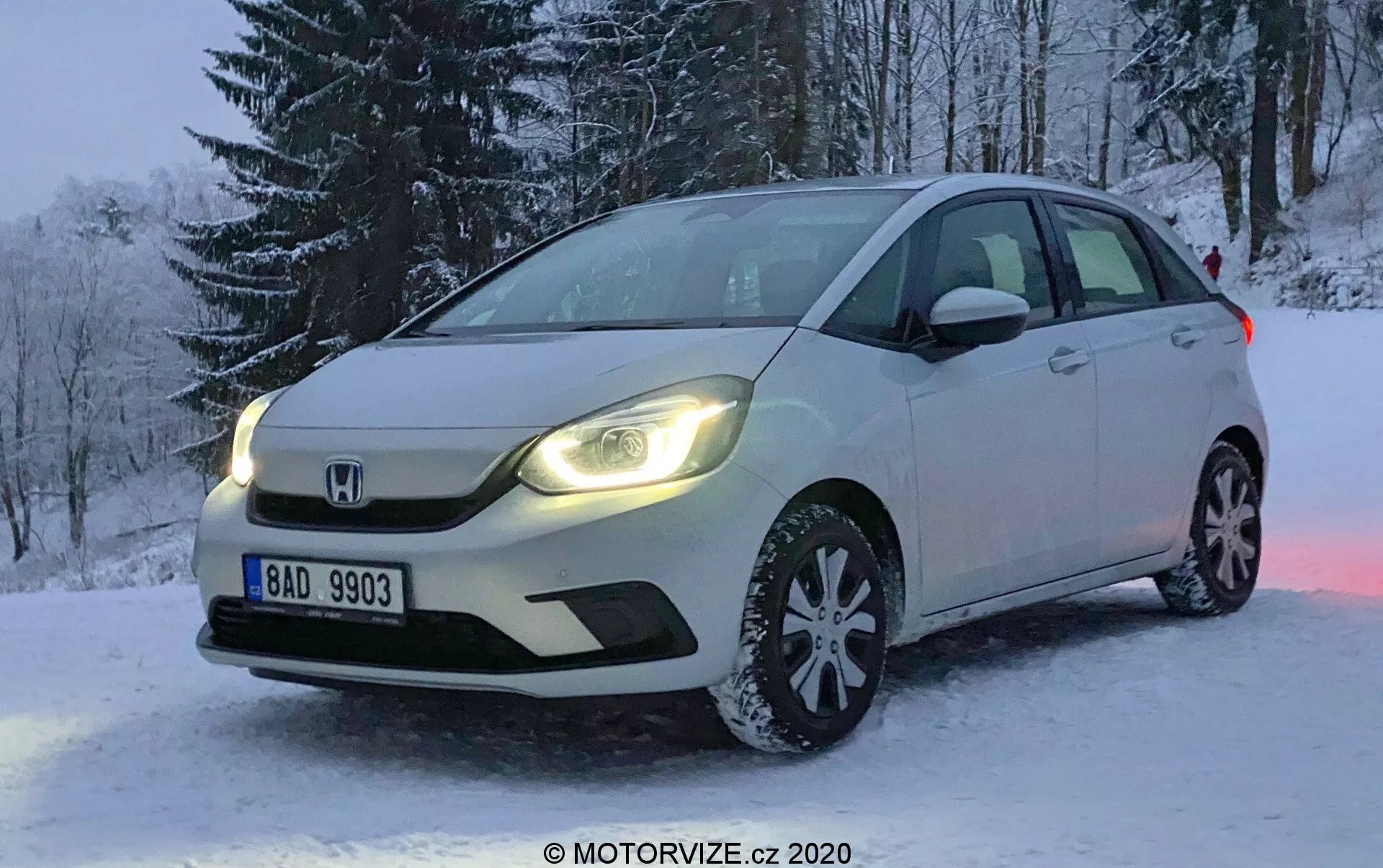 Profesjonalna fotografia białego Hondy Jazz z 2020 roku, zaparkowanego na zaśnieżonym terenie zimą. Zdjęcie wykonane jest pod kątem, skupiającym się na przedniej lewej stronie samochodu, ukazującym przedni grill, reflektory LED, maskę, lewy przedni wieloszprykowy aluminiowy felg z oponami zimowymi, przednią szybę i drzwi kierowcy. W tle widoczne są pokryte śniegiem drzewa, a tablica rejestracyjna jest czytelna. Przyciemnione światło sugeruje wczesny ranek lub późne popołudnie, z dziennymi światłami samochodu odbijającymi się od śniegu, podkreślając scenę. Pojazd posiada zaawansowane oświetlenie i zintegrowane kierunkowskazy w bocznych lusterkach, a charakterystyczny emblemat Honda wyraźnie widoczny jest na grillu.