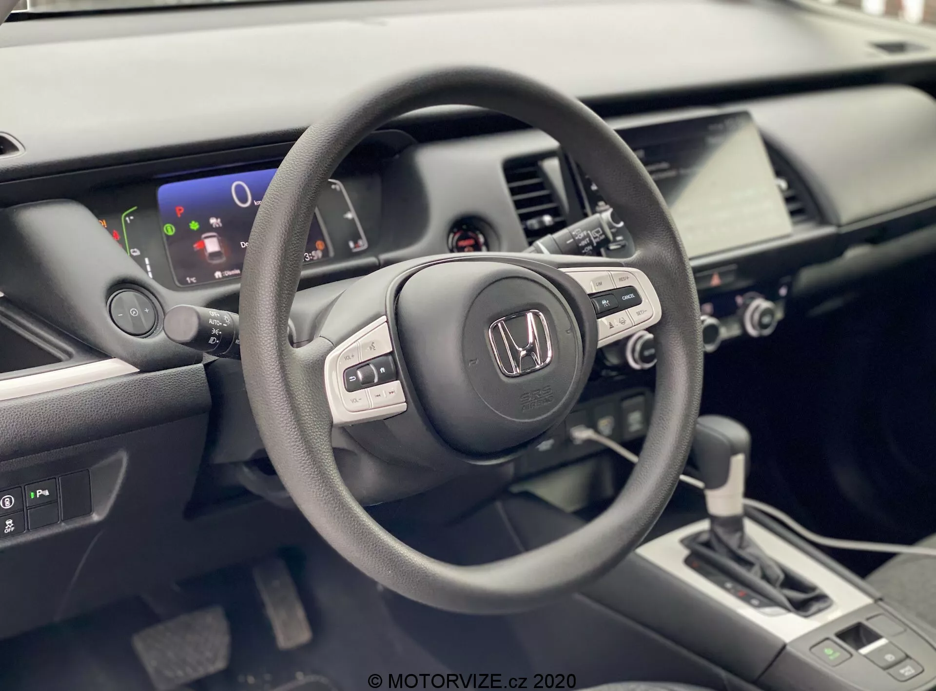Zdjęcie przedstawia wnętrze Hondy Jazz 2020 z perspektywy kierowcy. Skupia się na kierownicy z logo Honda, wyposażonej w kilka przycisków sugerujących zintegrowane funkcje audio, wyświetlacz informacji i być może tempomat. Za kierownicą widoczny jest częściowy wyświetlacz cyfrowy pokazujący wskaźnik poziomu naładowania baterii, wskazując, że pojazd może być hybrydą lub w pełni elektryczny. Dobrze oświetlone, nowoczesne wnętrze zawiera również konsolę centralną z dotykowym systemem infotainment, elementami sterującymi klimatyzacją i nowoczesną dźwignią zmiany biegów sugerującą automatyczną skrzynię biegów. Zdjęcie podkreśla matowe, satynowe i błyszczące wykończenia użytych materiałów w wnętrzu samochodu, co podkreśla ergonomiczny i bogaty w technologię design pojazdu.