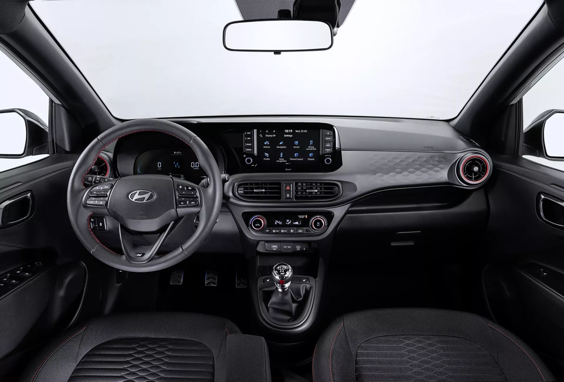 Wnętrze Hyundai i10 3. generacja lifting 2023 z nowoczesnym deskiem rozdzielczym, kierownicą trójramienną, zamontowanymi sterowaniami, systemem infotainment z ekranem dotykowym, kontrolą klimatu, okrągłymi nawiewami, automatyczną dźwignią zmiany biegów, wyrafinowanymi przednimi siedzeniami i eleganckim wnętrzem drzwi z oświetleniem ambientowym