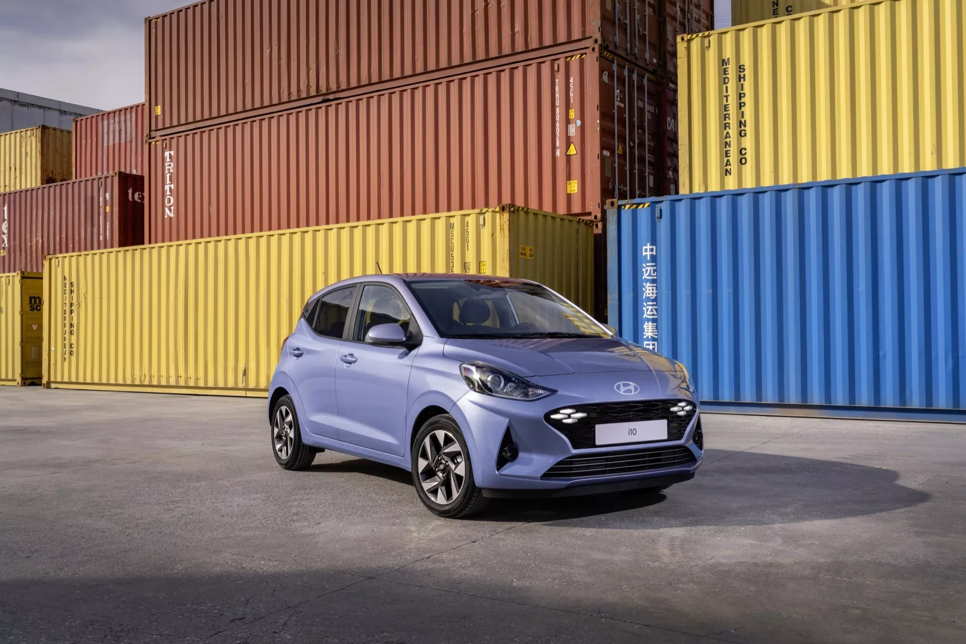 Model Hyundai i10 3. generacji lifting 2023 w dynamicznej pozie prezentujący reflektory LED, felgi aluminiowe i nowoczesne elementy wzornicze na kolorowym przemysłowym tle