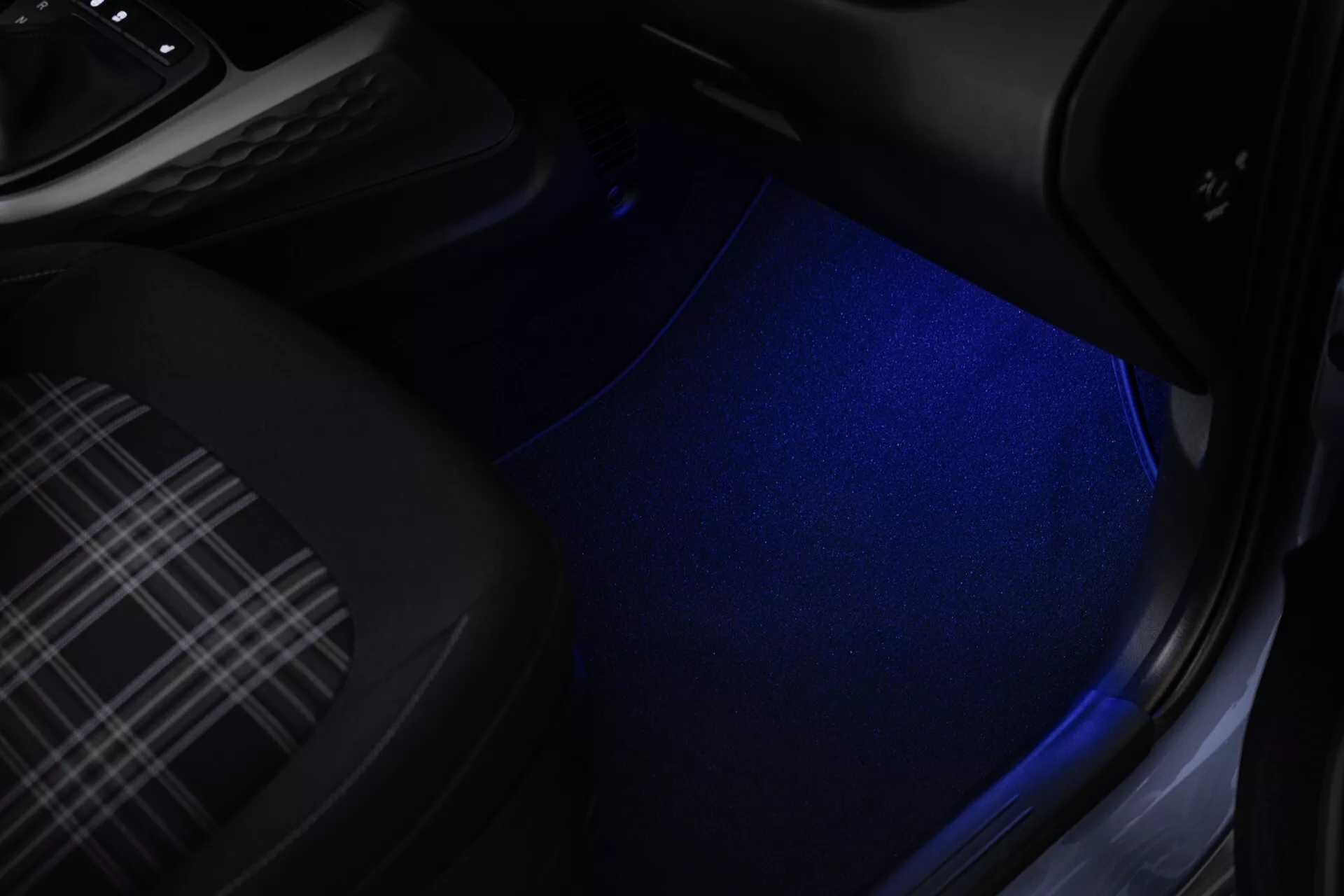 Wnętrze Hyundai i10 3. generacji po liftingu 2023 z podświetlaną przestrzenią na nogi oświetleniem LED w kolorze niebieskim, dywanikiem w kratę, częściowym widokiem na pedały automatycznej skrzyni biegów i progiem drzwi z potencjalnym elementem systemu dźwiękowego, podkreślające nowoczesne i stylowe elementy projektowe.