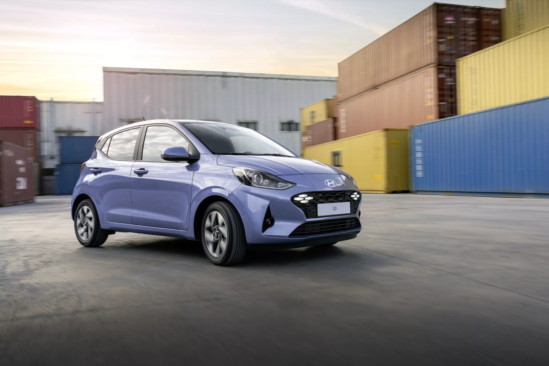Niebieski hatchback Hyundai i10 Facelift 2023 jedzie w strefie przemysłowej z kontenerami, z charakterystyczną atrapą, nowoczesnymi reflektorami, aerodynamicznym designem i fabrycznymi felgami aluminiowymi