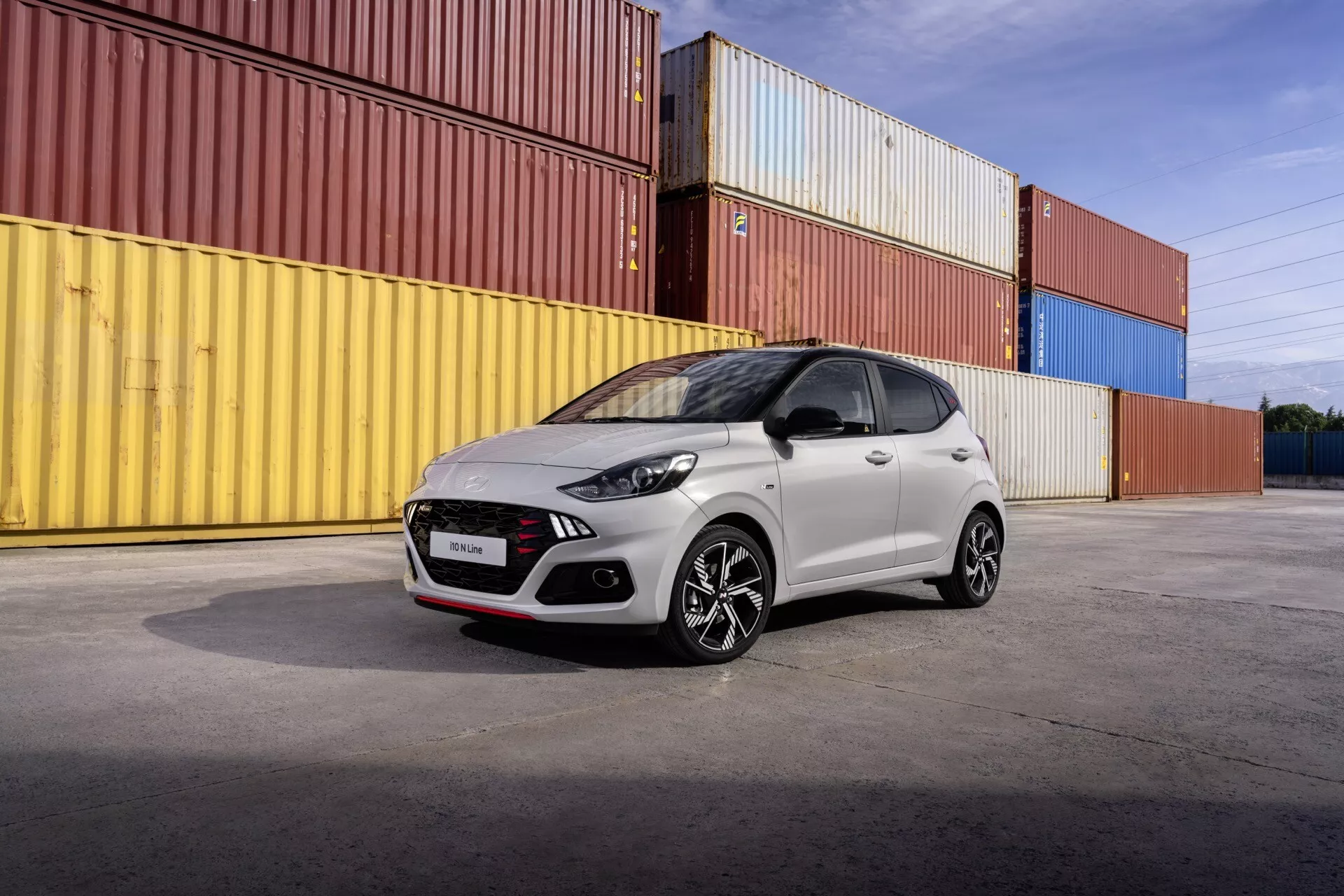Hyundai i10 3. generacji po liftingu, rok 2023, prezentowany na zewnątrz z eleganckim designem, charakterystycznymi reflektorami, grillem z centralnym emblematem, światłami przeciwmgielnymi i profil aerodynamiczny na tle kolorowych kontenerów