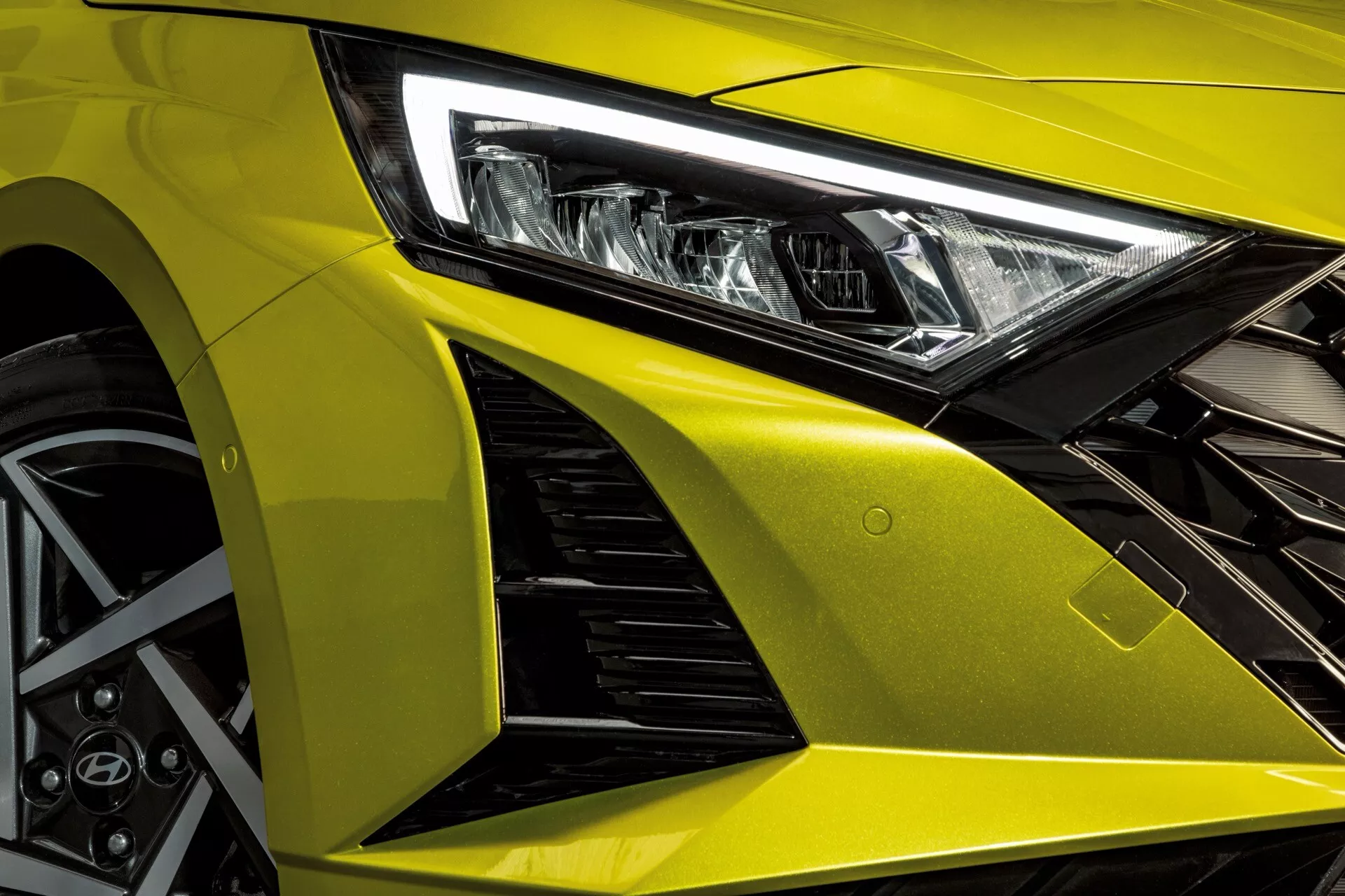 Widok z lewego przedniego kwartału Hyundai'a i20 2023, podkreślający agresywne linie projektowe, zaawansowany reflektor LED, metalicznie żółtą maskę, aerodynamiczny błotnik przedni z wlotami powietrza oraz sportowe koło stopowe z systemem hamulcowym o wysokiej wydajności