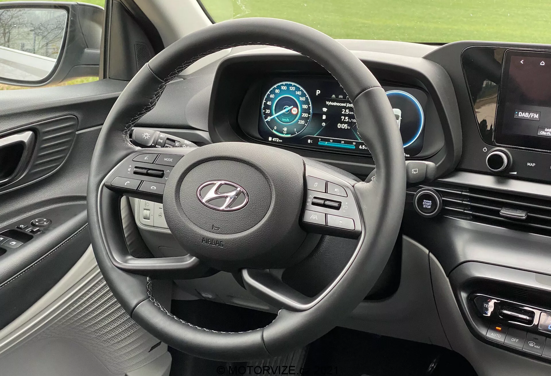 Widok wnętrza Hyundai i20 2020 z miejsca kierowcy, prezentujący trzyramienne koło kierownicy z wieloma przyciskami kontrolnymi, wyraźnie widoczne logo producenta, cyfrowy zestaw wskaźników w niebiesko-białej kolorystyce, prędkościomierz ustawiony na 0 km/h, pozycja zmiany biegów na Park, część konsoli centralnej z ekranem infotainment pokazującym mapę nawigacji, przyciski sterowania klimatem i ustawieniami pojazdu, miękka deska rozdzielcza z metalicznymi akcentami wokół otworów wentylacyjnych i kierownicy oraz częściowy widok otworu wentylacyjnego po stronie pasażera.