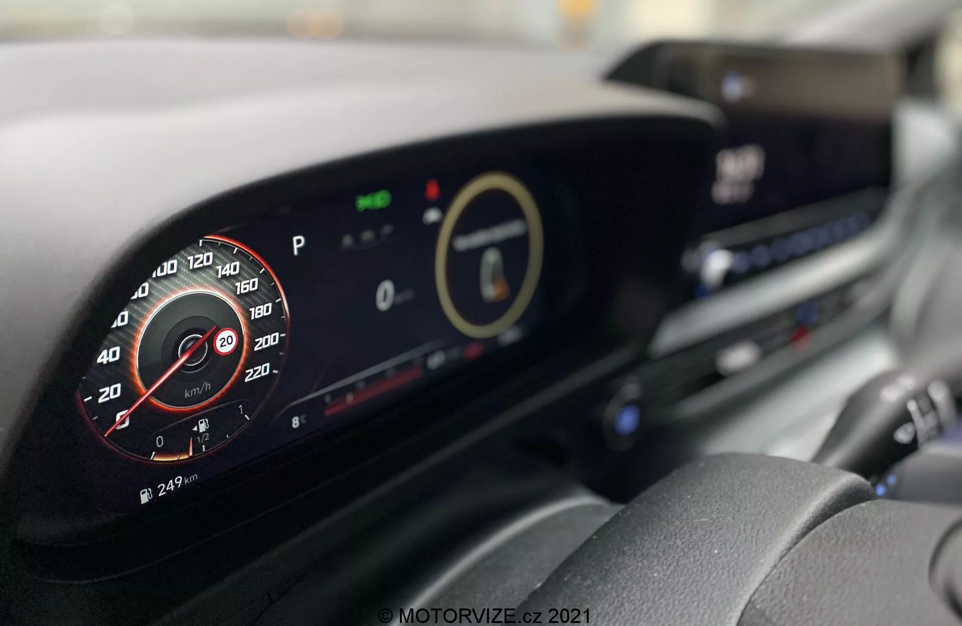Wnętrze Hyundai i20 2020 z nowoczesną deską rozdzielczą. Zdjęcie wykonane z fotela kierowcy, skupiające się na cyfrowym zestawie wskaźników i centralnym wyświetlaczu, prezentujące szczegóły, takie jak symulowany analogowy obrotomierz, wskaźniki ekologiczne oraz system info-rozrywkowy. Widoczne są także dodatkowe funkcje, takie jak kratki wentylacyjne i pokrętła sterowania klimatyzacją.