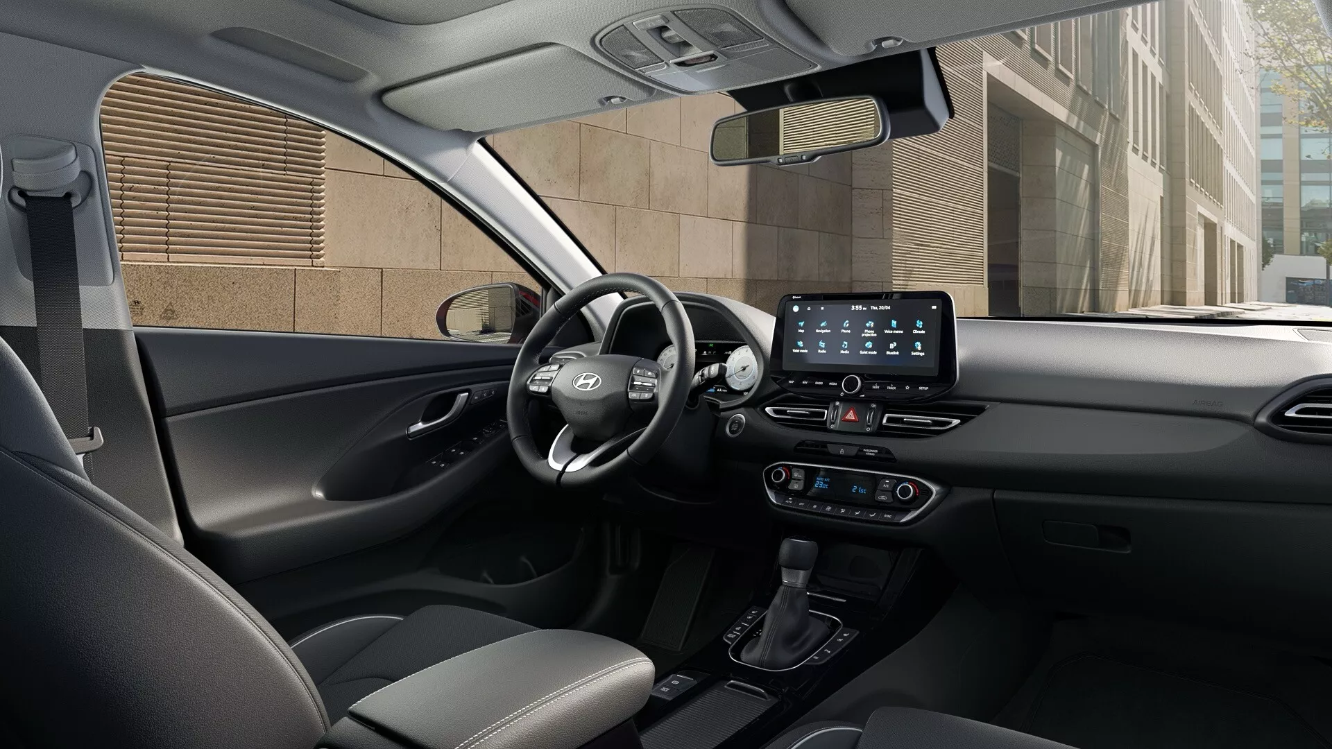 Widok wnętrza Hyundai i30 2024 (3. generacja, facelifting 3) z tylnego siedzenia, ukazujący deskę rozdzielczą, konsolę środkową i strefę kierowcy. Kierownica po lewej stronie ma przyciski wielofunkcyjne. Cyfrowy wyświetlacz za kierownicą pokazuje informacje o jazdzie. Konsola środkowa zawiera dotykowy system infotainment z funkcjami łączności, przełącznikami klimatyzacji i automatyczną dźwignię zmiany biegów. Siedzenia i wnętrza drzwi są pokryte ciemną tkaniną lub skórą z dwukolorowym wzorem. Układ podkreśla technologię i komfort z zaawansowanymi systemami wspomagania kierowcy, łącznością Bluetooth lub USB oraz dźwiękiem premium. Widoczne są boczne szyby i lusterka z możliwymi elektronicznymi kontrolkami.