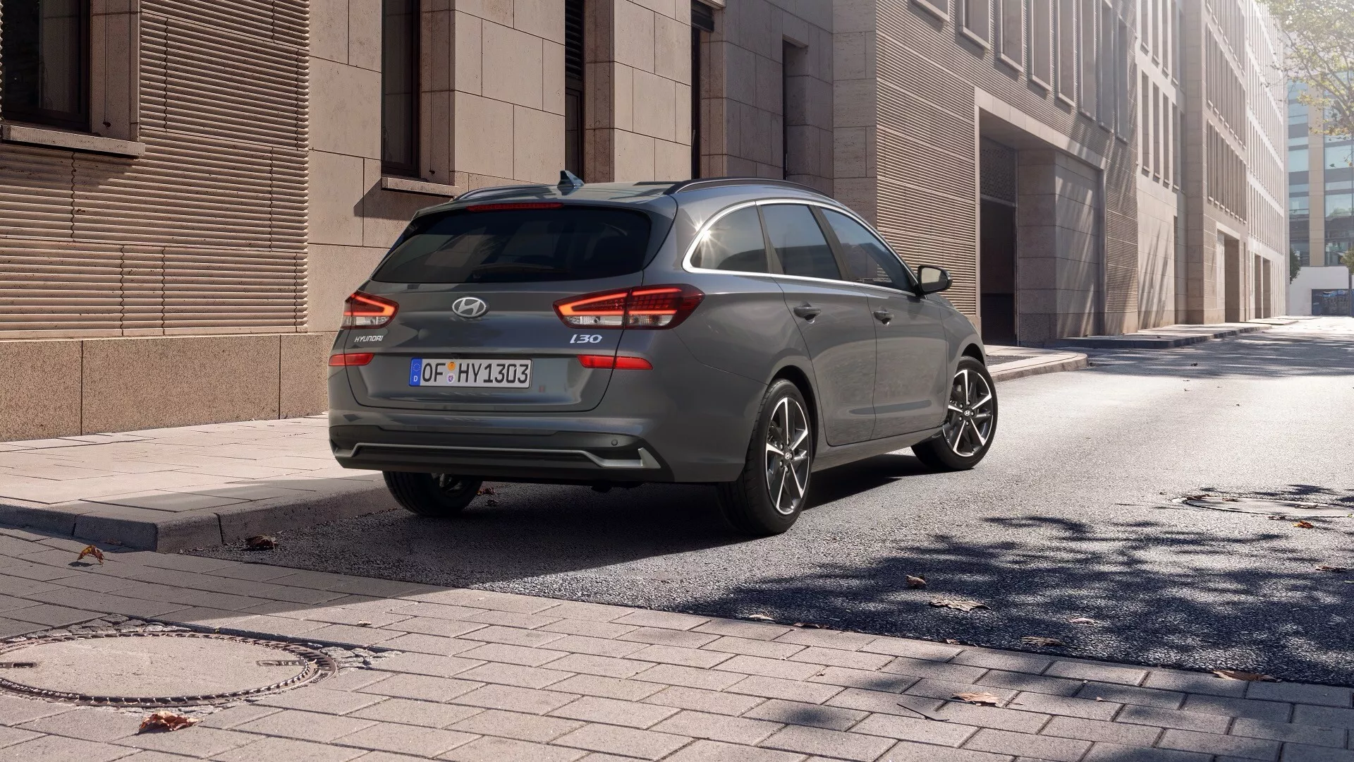 Widok z tyłu pod kątem trzech czwartych na szarego Hyundai i30 2024 typu kombi, pokazujący bagażnik typu hatchback, tylną szybę, stronę pasażera, nowoczesne tylne światła i wieloramienne felgi aluminiowe, zaparkowanego na ulicy miasta pod jasnym niebem.