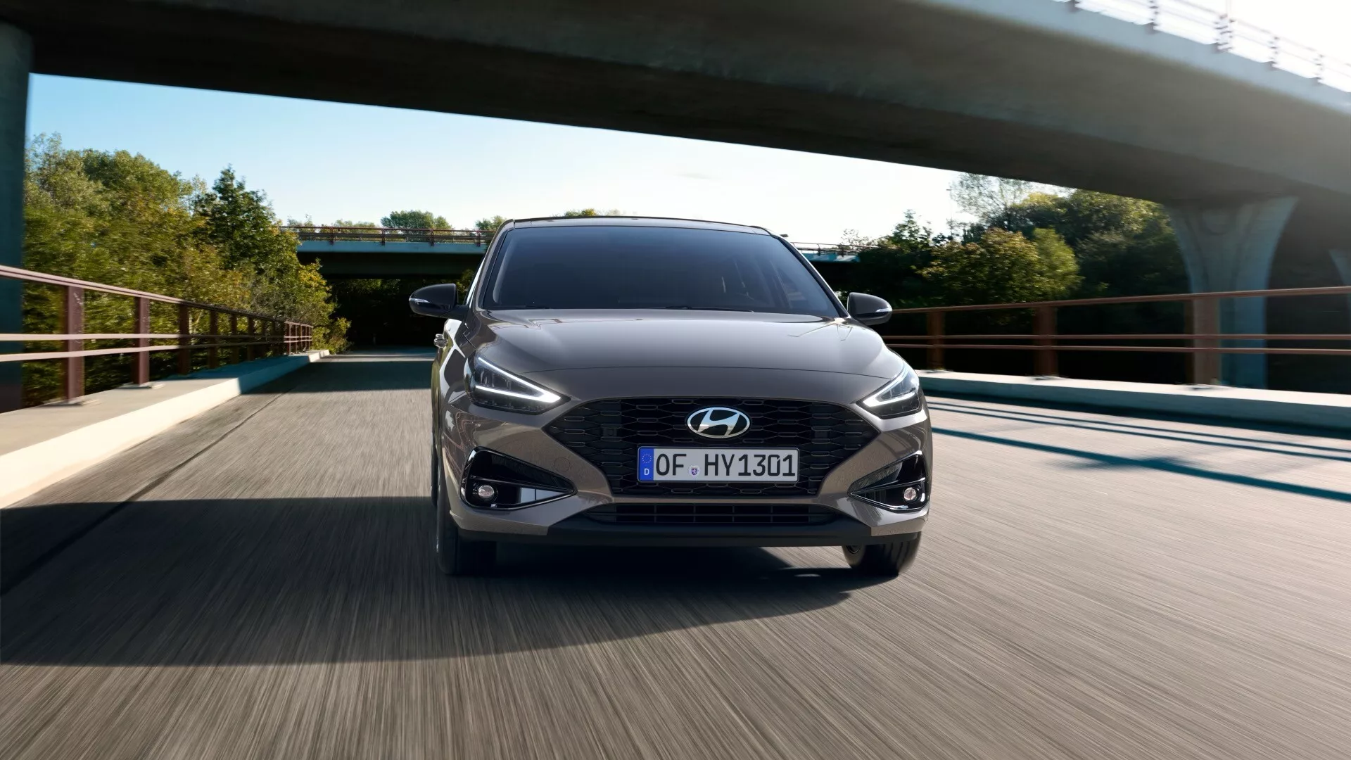 Ujęcie centralne przodu Hyundai i30 2024 (trzecia generacja po liftu) uchwycone w ruchu. Kratka grilla ma duży wzór siatki z emblematem Hyundai pośrodku. Eleganckie reflektory LED zakrzywiają się do tyłu w kierunku błotników, z reflektorami przeciwmgielnymi poniżej. Pojazd ma tablicę rejestracyjną w stylu europejskim. Rozmycie ruchu kół i drogi wskazuje na średnią do wysoką prędkość. Ujęcie pod wiadukt em z jasnym niebem i drzewami w tle, podkreśla nowoczesny design i technologiczne funkcje samochodu, takie jak adaptacyjne reflektory LED i wieloramienne felgi aluminiowe.