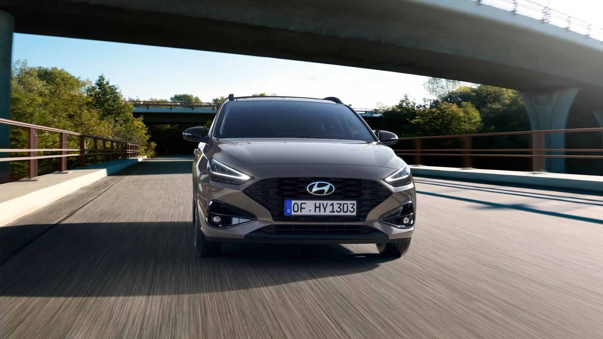 Obraz Hyundai i30 2024 (3. generacja - lifting 3) uchwycony z przedniego niskiego kąta podczas jazdy po drodze. Widok z dołu podkreśla przedni grill, zderzak i dolną część nadwozia, z włączonymi reflektorami LED i dobrze widocznym logo Hyundai. Grill ma charakterystyczny wzór siatki, z tablicą rejestracyjną w europejskim stylu. Dynamiczne linie konturowe na masce opadają w kierunku grilla, poprawiając aerodynamiczny wygląd. Połyskujący ciemny metalik, łagodne oświetlenie oraz nowoczesne otoczenie infrastruktury z mostem sugerują, że jest późne popołudnie lub wczesny poranek w obszarze miejskim lub podmiejskim.