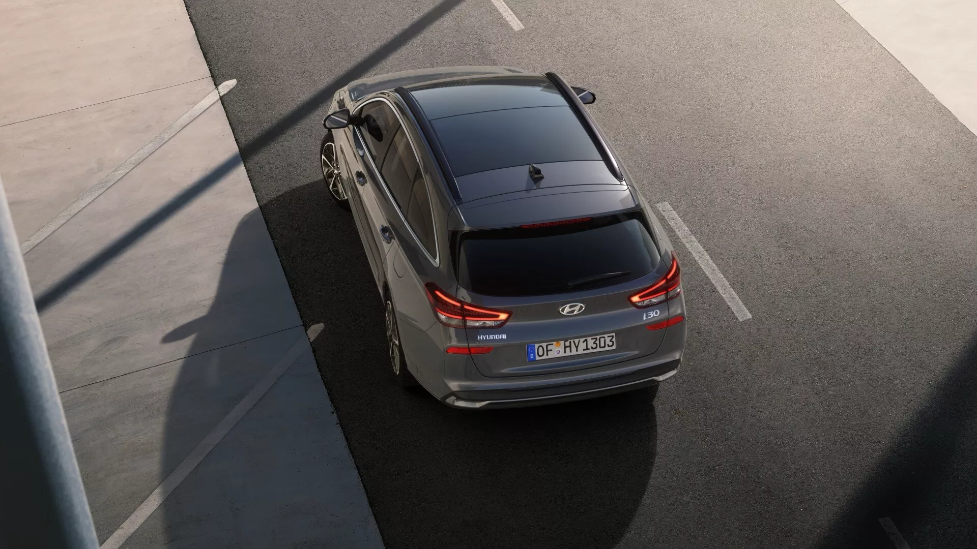 Widok z dużej wysokości na tylną i górną część Hyundai i30 2024, wyróżniający wydłużoną tylną część nowoczesnego hatchbacka. Charakterystyczne cechy to tylna szyba wyposażona w odmrażacz z wycieraczką, spoiler na górze tylnej klapy dla aerodynamicznego wyglądu, szczegółowe tylne światła zwiększające widoczność, centralnie umieszczona tablica rejestracyjna, emblemat marki i oznaczenia modelu na bagażniku, oraz odblaskowe paski na tylnym zderzaku poprawiające widoczność przy słabym oświetleniu.