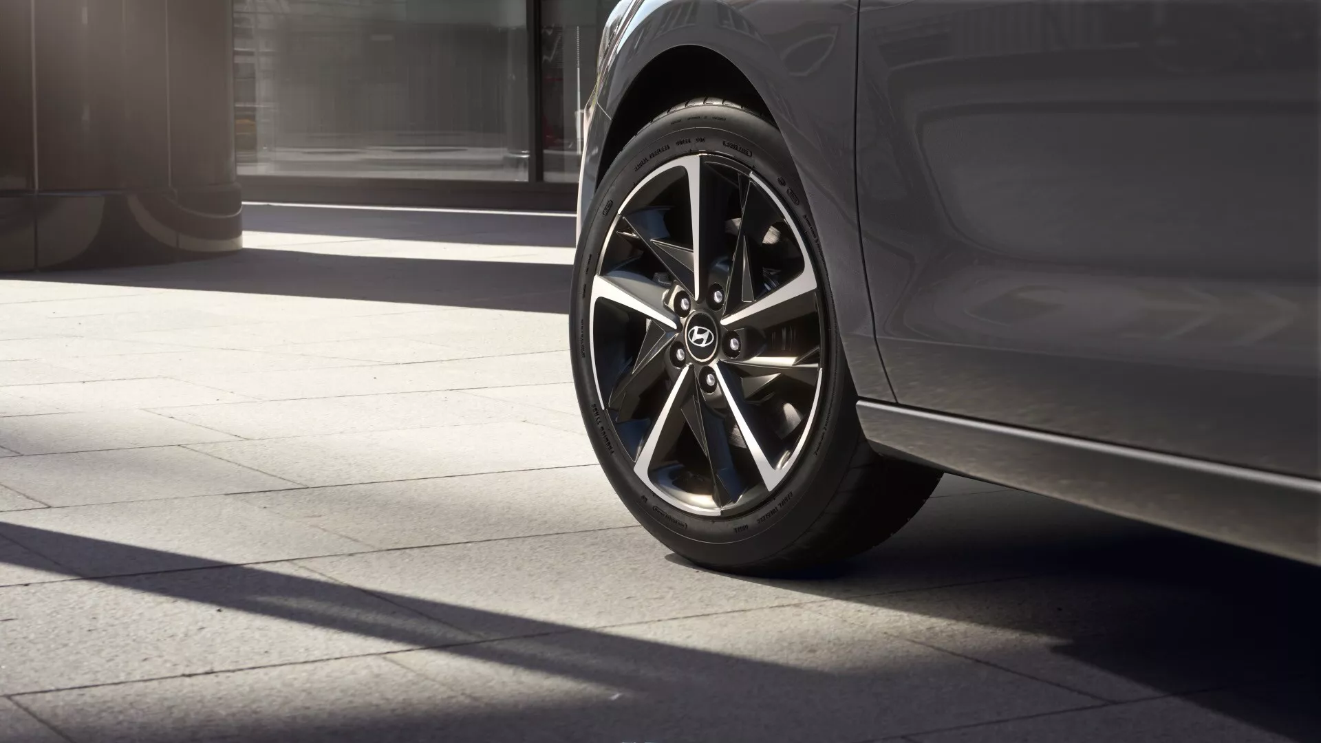 Zbliżenie na przednie lewe koło i część drzwi kierowcy w Hyundai i30 2024 (lifting trzeciej generacji). Ujęcie z niskiego kąta podkreśla nowoczesną, wieloramienną felgę ze stopu metali z ciemnym metalicznym wykończeniem, zestawioną z niskoprofilową oponą sportową. Podkreślony jest elegancki, minimalistyczny design dolnej części drzwi i wpuszczony uchwyt. Światło słoneczne rzuca wyraźne cienie na chodnik, sugerując środek dnia lub wczesne popołudnie.