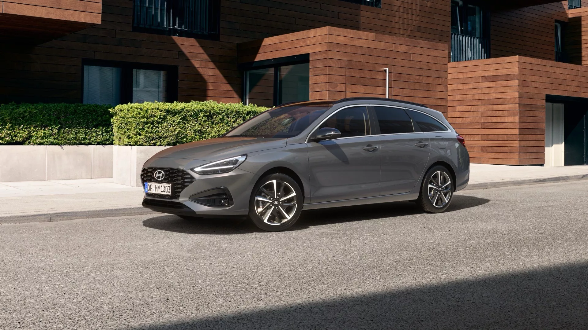 Obrazek przedstawia Hyundai i30 2024 (3. generacja facelift) uchwycony z niskiego kąta czołowego trzech czwartych, podkreślając zarówno przednie, jak i boczne cechy designu. Widok z przodu pokazuje skomplikowane reflektory LED z dziennymi światłami, kratkę specyficzną dla marki z centralnie umieszczonym logo oraz zderzak z wlotami powietrza do chłodzenia silnika i aerodynamiki. Widok z boku ukazuje felgi aluminiowe, aerodynamiczne lusterka boczne z zintegrowanymi kierunkowskazami oraz dynamiczną linię charakterystyczną biegnącą wzdłuż całej długości pojazdu. Styl nadwozia wydaje się być hatchbackiem lub kombi, z widocznymi tylnymi drzwiami i prawdopodobnie szyberdachem lub dachem panoramicznym. Samochód jest zaparkowany na ulicy, co wskazuje na jego konwencjonalny rozmiar odpowiedni do środowisk miejskich. Ogólnie rzecz biorąc, design jest nowoczesny i elegancki, sugerujący zaawansowane funkcje i wybierany przez praktyczne osoby lub rodziny preferujące nowoczesne udogodnienia i styl.