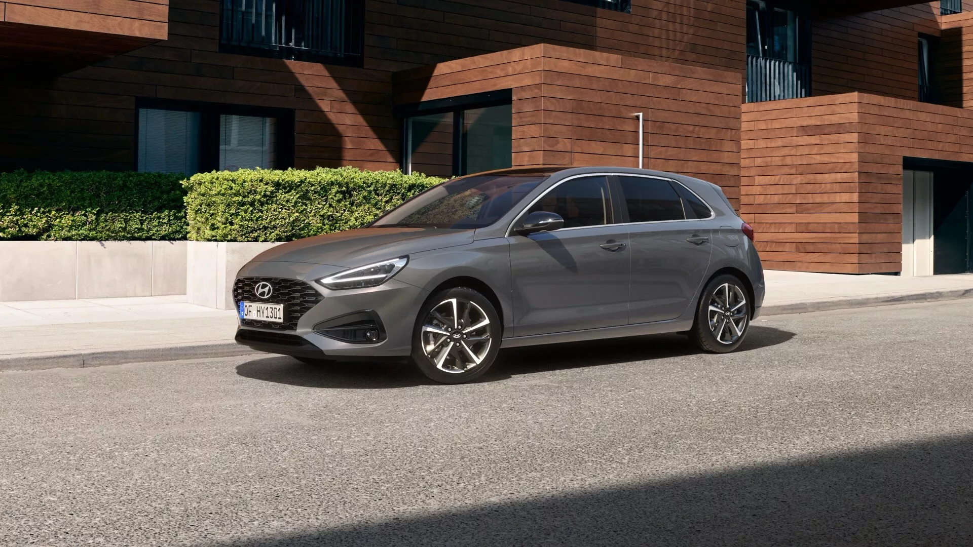Hyundai i30 2024 sfotografowany pod lekko podwyższonym kątem, ukazując jego ogólny design, dach i część maski. Samochód jest zaparkowany na osiedlu mieszkaniowym z nowoczesną architekturą w tle, prezentując czterodrzwiowy hatchback z felgami ze stopów lekkich, co sugeruje średni lub wyższy poziom wykończenia. W pełni widoczny jest boczny profil od strony kierowcy, podkreślający jego aerodynamiczny kształt, przyciemniane szyby i antenę na dachu, z otoczeniem zapewniającym wyraźny widok na jego stylowy, nowoczesny design.