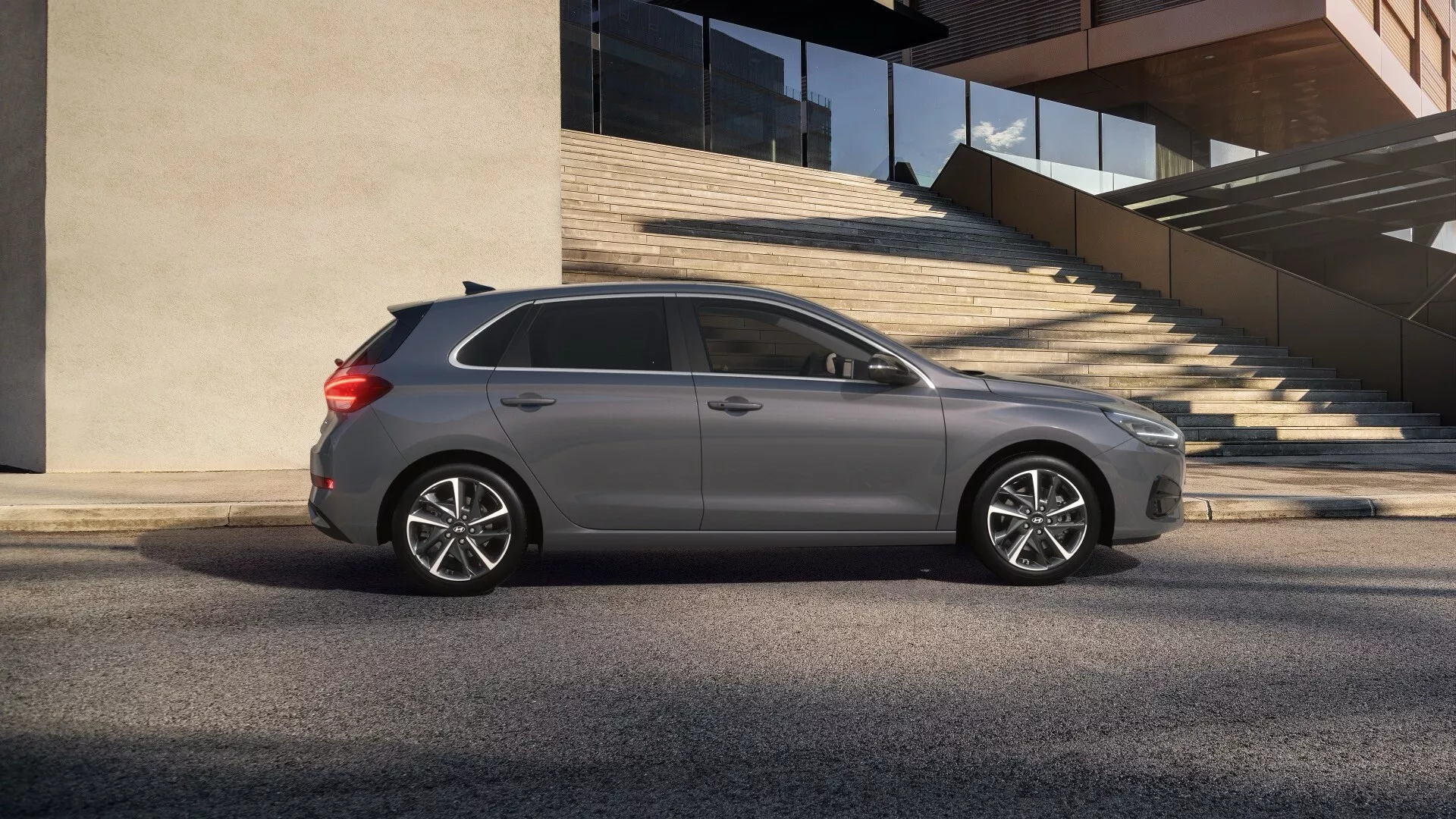 Profesjonalne zdjęcie bocznego profilu Hyundai i30 2024 (3. generacja facelifting) szarego hatchbacka uchwycone pod niskim kątem na miejskiej ulicy, podkreślające nowoczesny design reflektorów, część przedniego grilla, eleganckie felgi aluminiowe i wyrafinowane linie nadwozia, koncentrując się na jego przydatności do jazdy po mieście.