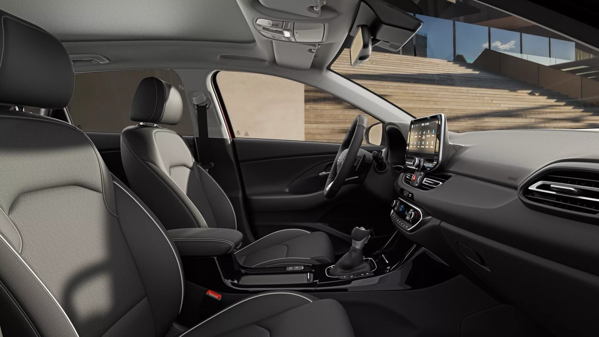 Widok wnętrza Hyundai i30 2024, trzecia generacja facelift. Przednia część kabiny widziana jest z nieco wysokiego kąta, patrząc na skos w kierunku strony kierowcy. Kierownica ma zintegrowane przyciski do sterowania multimediami, tempomatem i komendami głosowymi. Za kierownicą znajduje się zestaw wskaźników, który może być cyfrowym lub częściowo cyfrowym wyświetlaczem. Na konsoli środkowej znajduje się wyraźny dotykowy system informacyjno-rozrywkowy do nawigacji, odtwarzania multimediów i ustawień samochodu. Poniżej znajdują się ustawienia klimatyzacji dwustrefowej, automatyczny przełącznik biegów, uchwyty na kubki i miejsce do przechowywania. W pobliżu dźwigni zmiany biegów widoczny jest port USB. Siedziska wydają się być skórzane z kontrastowymi szwami, co wskazuje na nacisk na wygodę i styl. Widać nowoczesne funkcje łączności, wygody i interfejsu użytkownika, z naciskiem na sterowanie dotykiem i komendy głosowe.