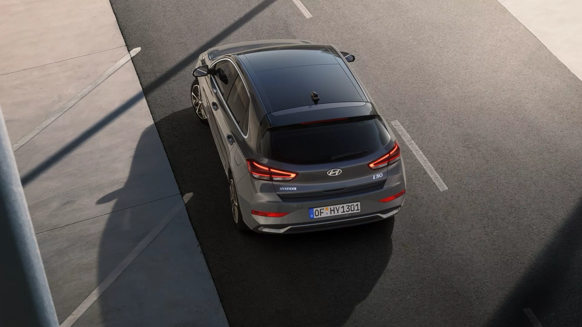 Widok z góry na tyl z lotu ptaka na SUV Hyundai IONIQ 5, podkreślający nowoczesny, opływowy design w jasnym kolorze, prawdopodobnie srebrnym lub jasnoszarym. Wyraźny obraz ukazuje tylne elementy, w tym tylne światła, tylną wycieraczkę, spoiler, emblemat Hyundai, tablicę rejestracyjną, tylne czujniki lub kamery oraz antenę na dachu. Częściowy widok górnej strony i kół z nowoczesnym projektem ze stopu metali lekkich. Samochód wydaje się być na podwyższonej drodze lub moście bez otaczającego ruchu, co sugeruje, że może być w ruchu lub zaparkowany. Ujęcie, wykonane pod wysokim kątem w dobrych warunkach oświetleniowych, prawdopodobnie rano lub po południu, podkreśla zaawansowaną technologię i estetykę pojazdu, prawdopodobnie do celów promocyjnych.