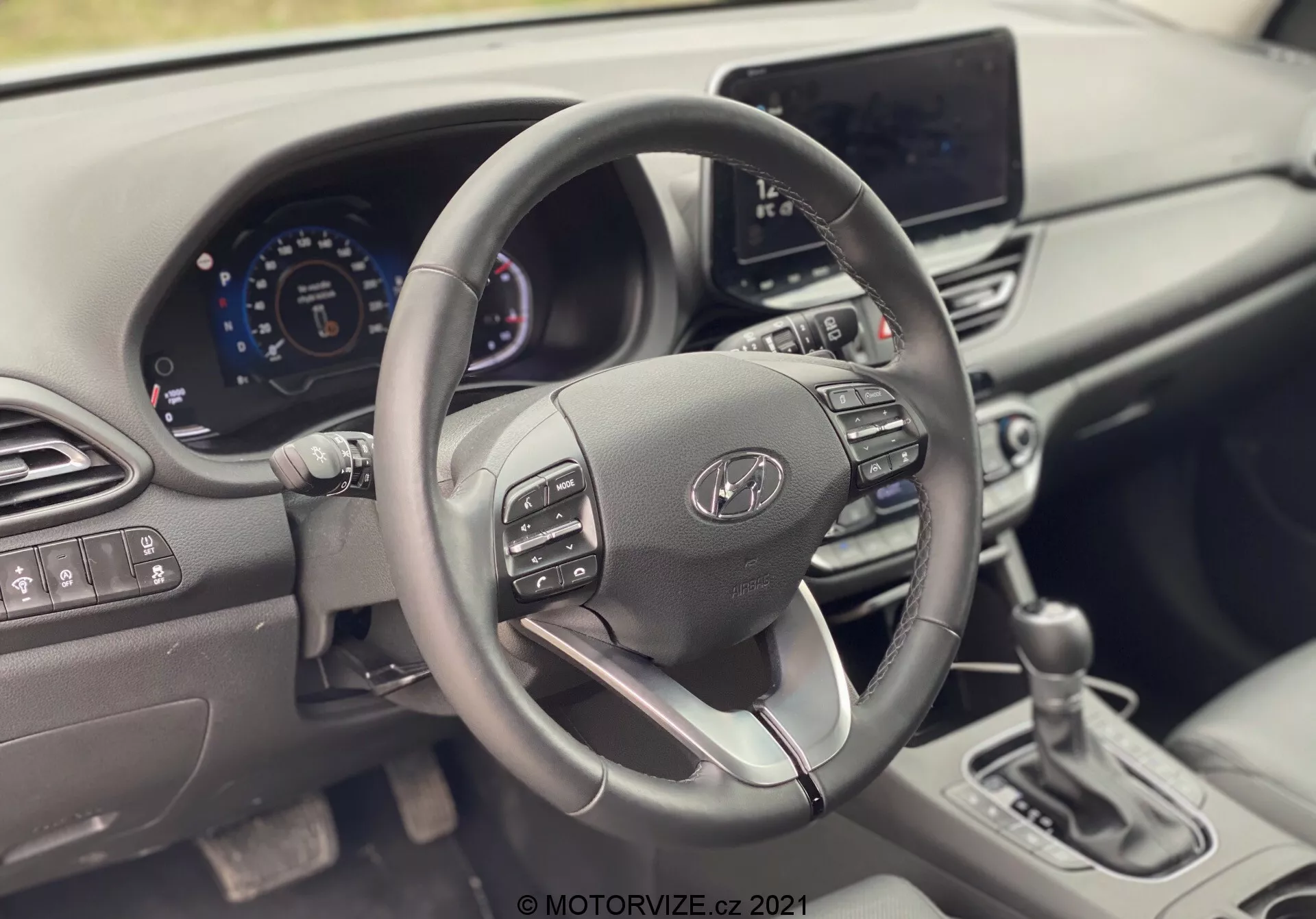 Widok wnętrza Hyundai i30 Kombi 2020 skupiający się na kierownicy i desce rozdzielczej. Zdjęcie wykonane z miejsca pasażera, pokazujące elegancką kierownicę z przyciskami sterowania, cyfrowy zestaw wskaźników i system infotainment. Nowoczesny design obejmuje kierownicę pokrytą skórą z dolnym ramieniem w kolorze srebrnym, nawiewy i minimalną liczbę fizycznych przycisków, podkreślające zaawansowane technologie motoryzacyjne i ergonomiczny design.