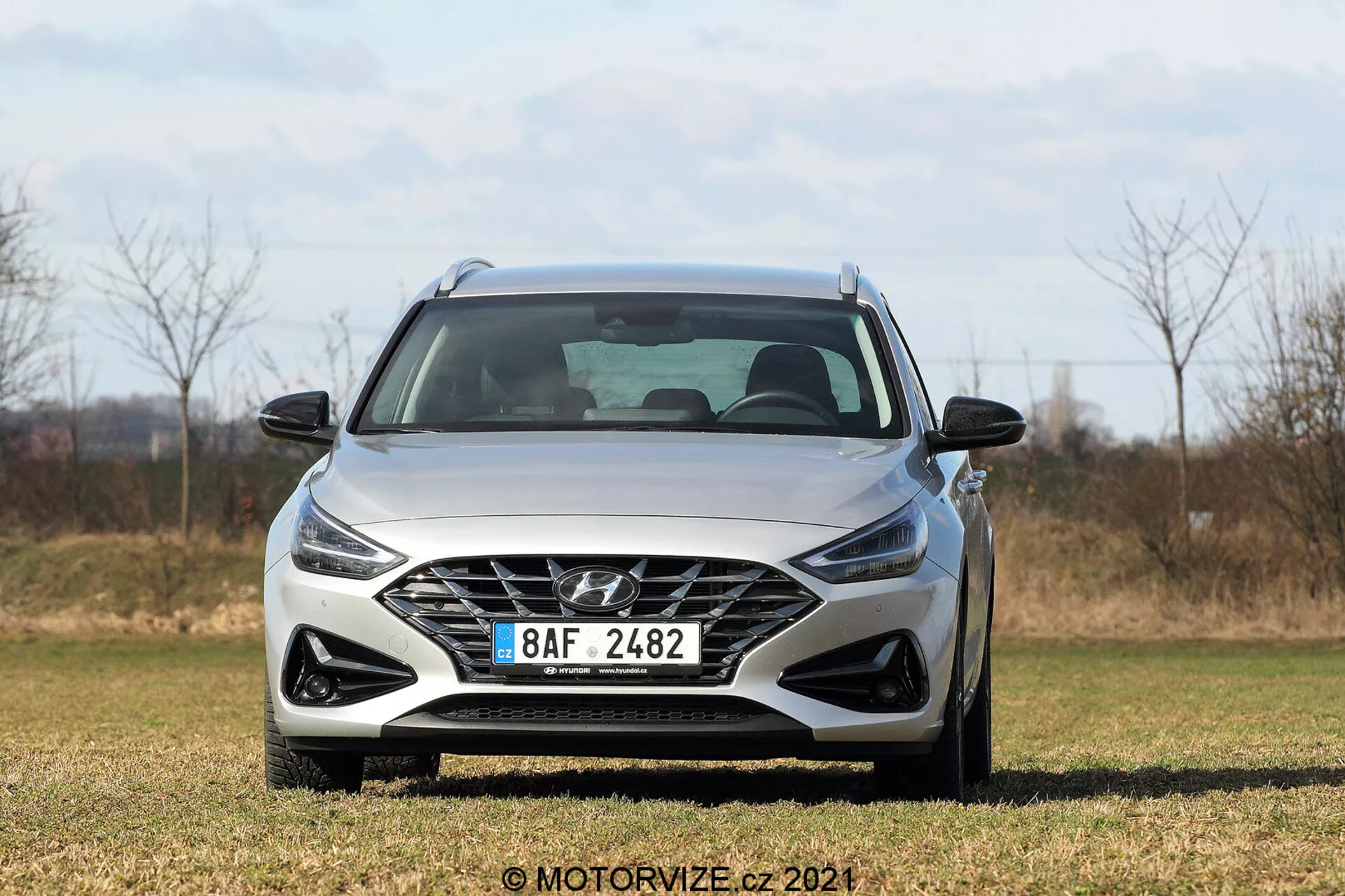 Widok z przedniego kąta na Hyundai i30 Kombi 2020, prezentujący wielokątną kratkę z logo producenta, ostre kątowe reflektory, rzeźbiony przedni zderzak, subtelne linie maski i lusterka boczne na obszarze trawiastym z naturalnym tłem.